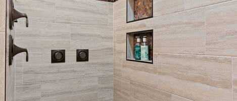Zona doccia con soffione a pioggia, doccetta manuale e ripiano con articoli da toilette.