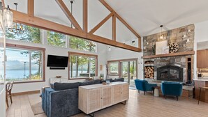 A spacious living area with a large sectional sofa, a stone fireplace, and a view of a lake. - Beautiful Waterfront Cabin with a Hot Tub & Sauna (Nordman)