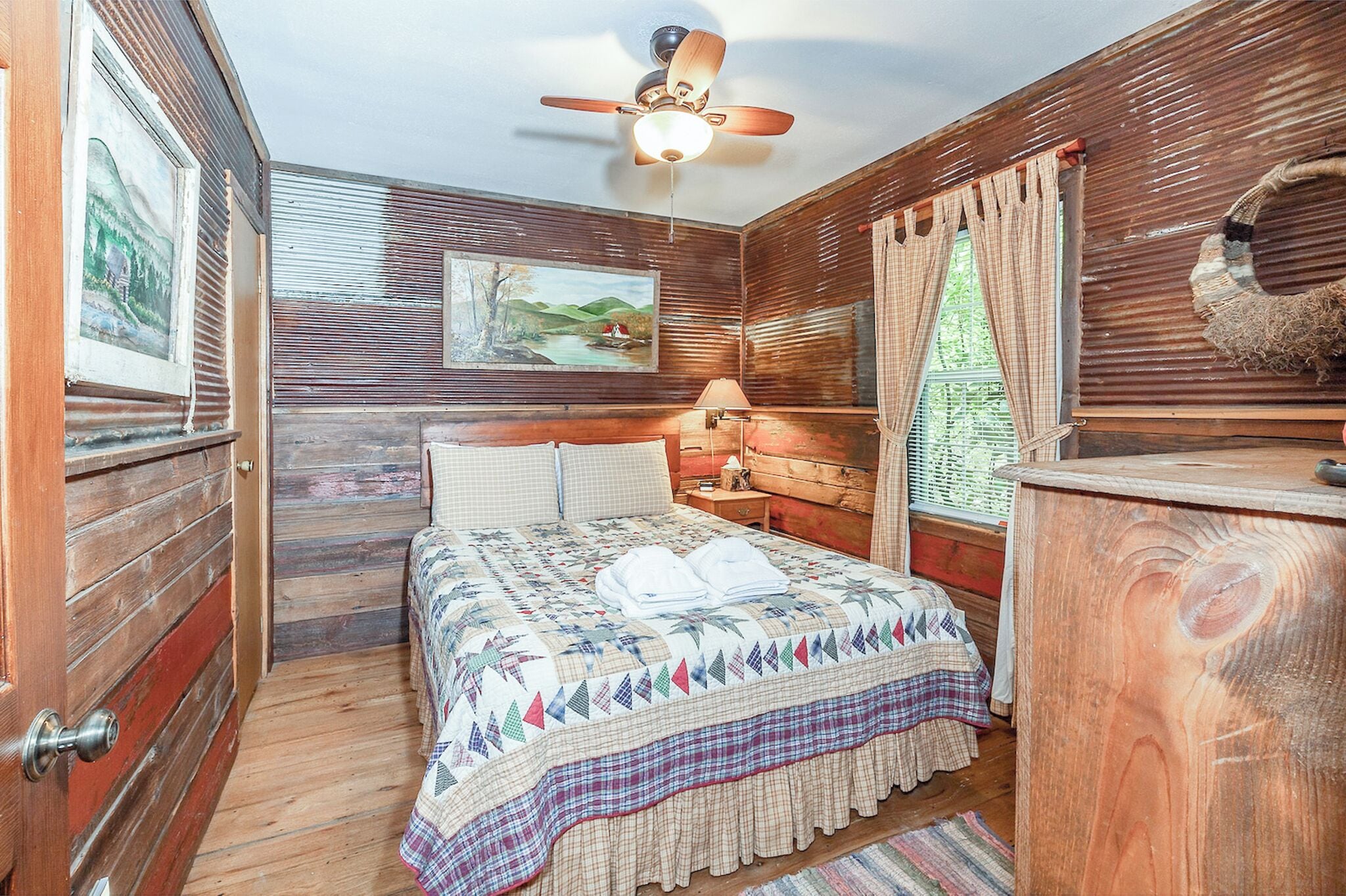 Een slaapkamer in chaletstijl met een bed, een plafondventilator, een schilderij en een raam met gordijnen.