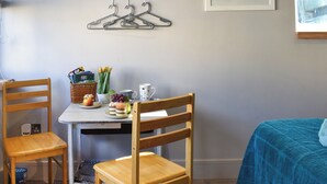 A room with a desk, two wooden chairs, a bed with a blue cover, a wall-mounted fish-shaped hanger rack, a framed painting, and a basket with books and tulips.