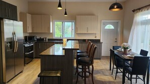 Cozinha moderna com ilha central, armários escuros, eletrodomésticos de aço inoxidável e área de jantar com cadeiras pretas e uma mesa.