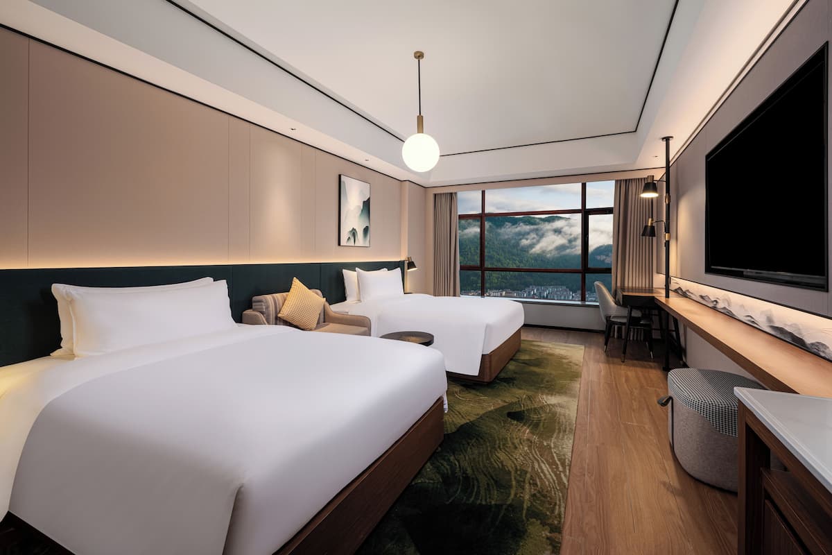 A modern hotel room with two beds, a desk, a large TV, and a view of mountains.
