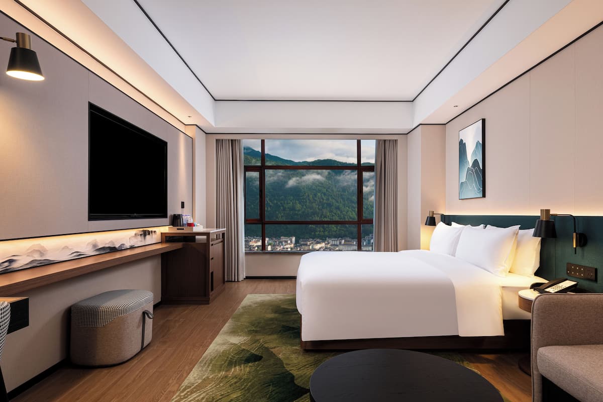 A modern hotel room with a large bed, a desk, a flat-screen TV, a view of mountains, and a cityscape through the window.