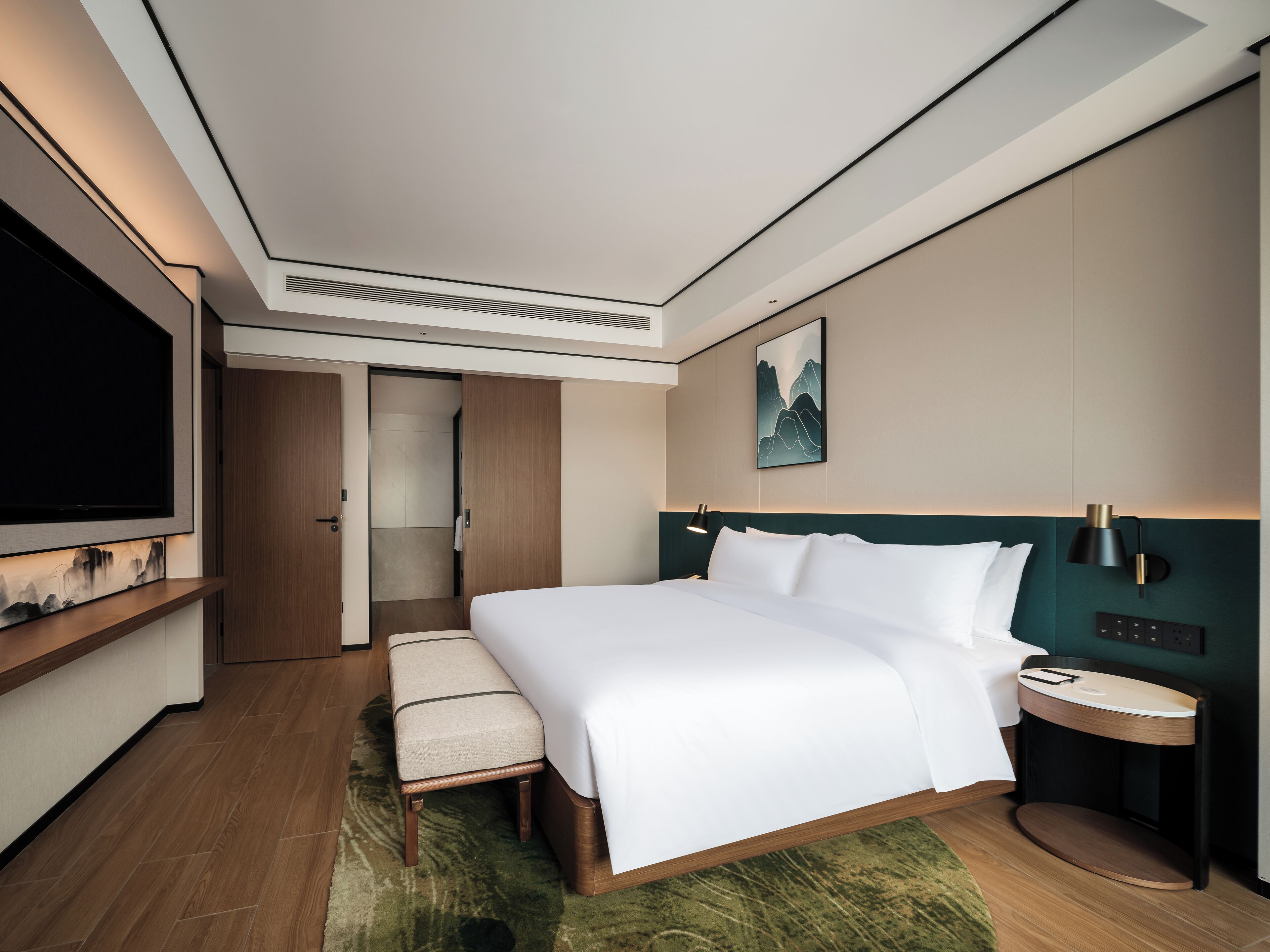A modern hotel room with a large bed, a bedside table, a TV mounted on the wall, and a door leading to a bathroom.