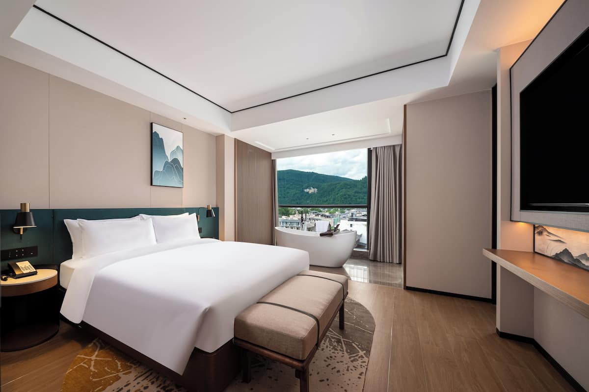 A modern hotel room with a large bed, a sitting area, a bathtub, and a TV.