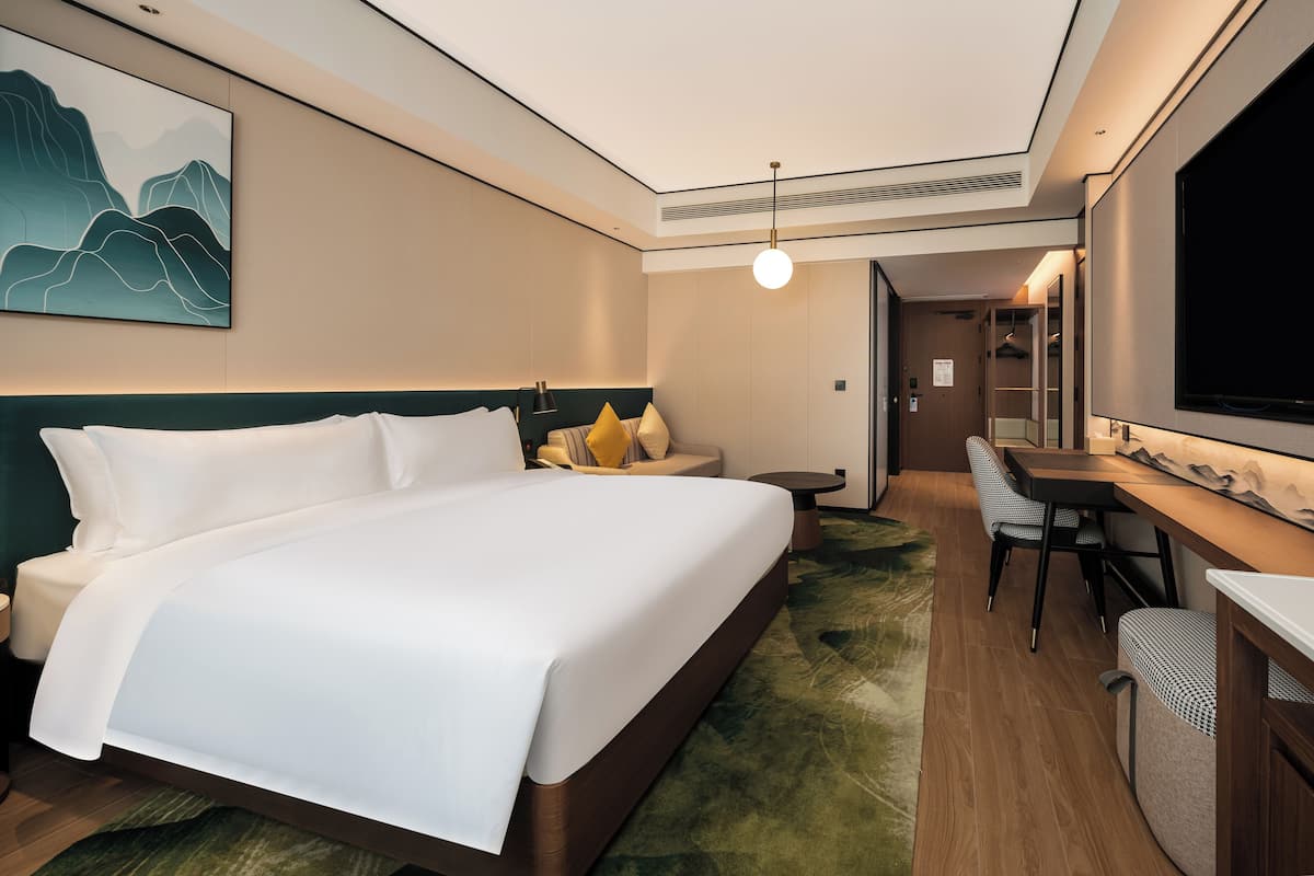 A modern hotel room with a large bed, a desk, a chair, a television, and a painting on the wall.