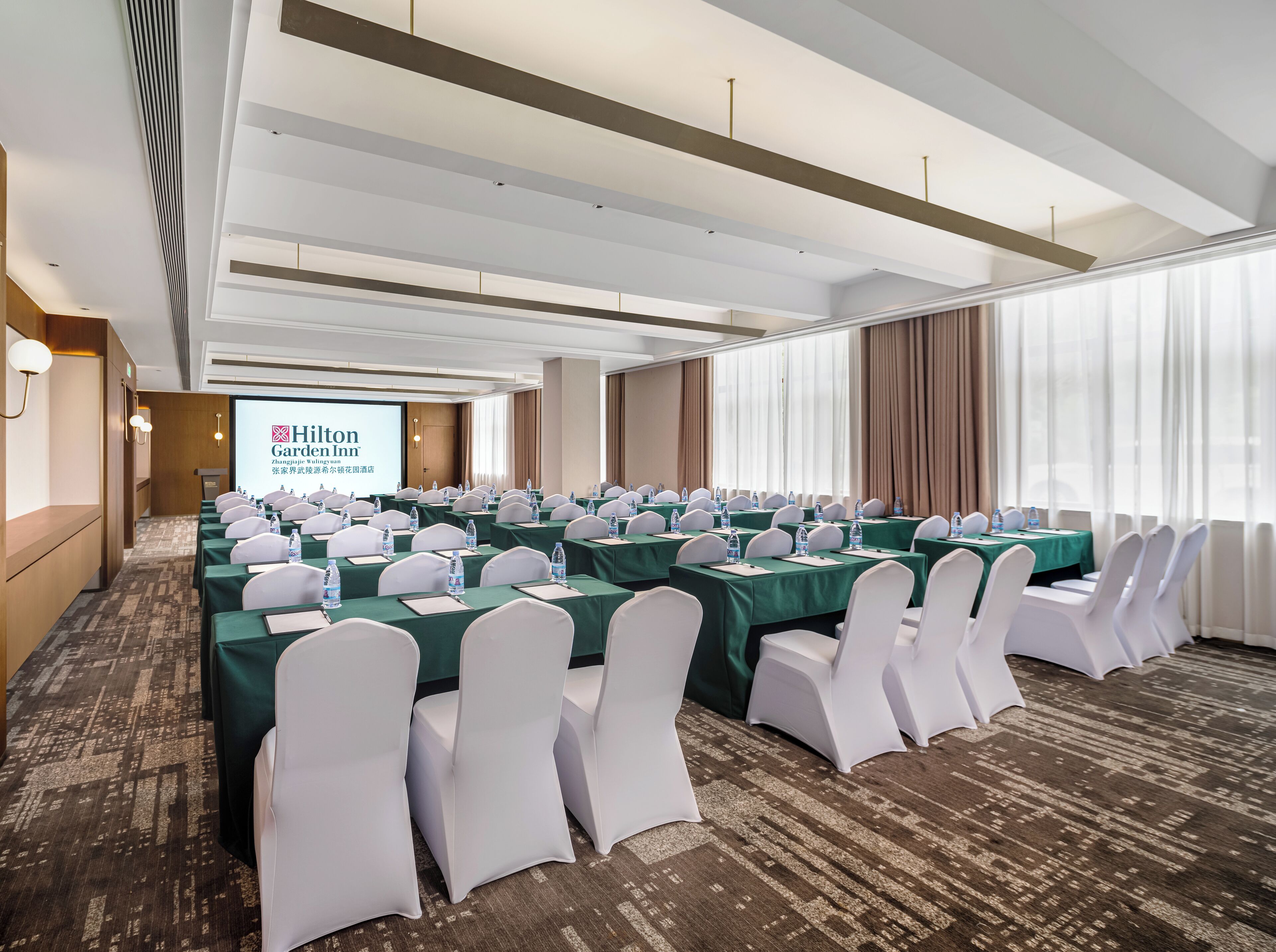 A conference room with rows of tables, chairs, and a projection screen displaying Hilton Garden Inn branding.