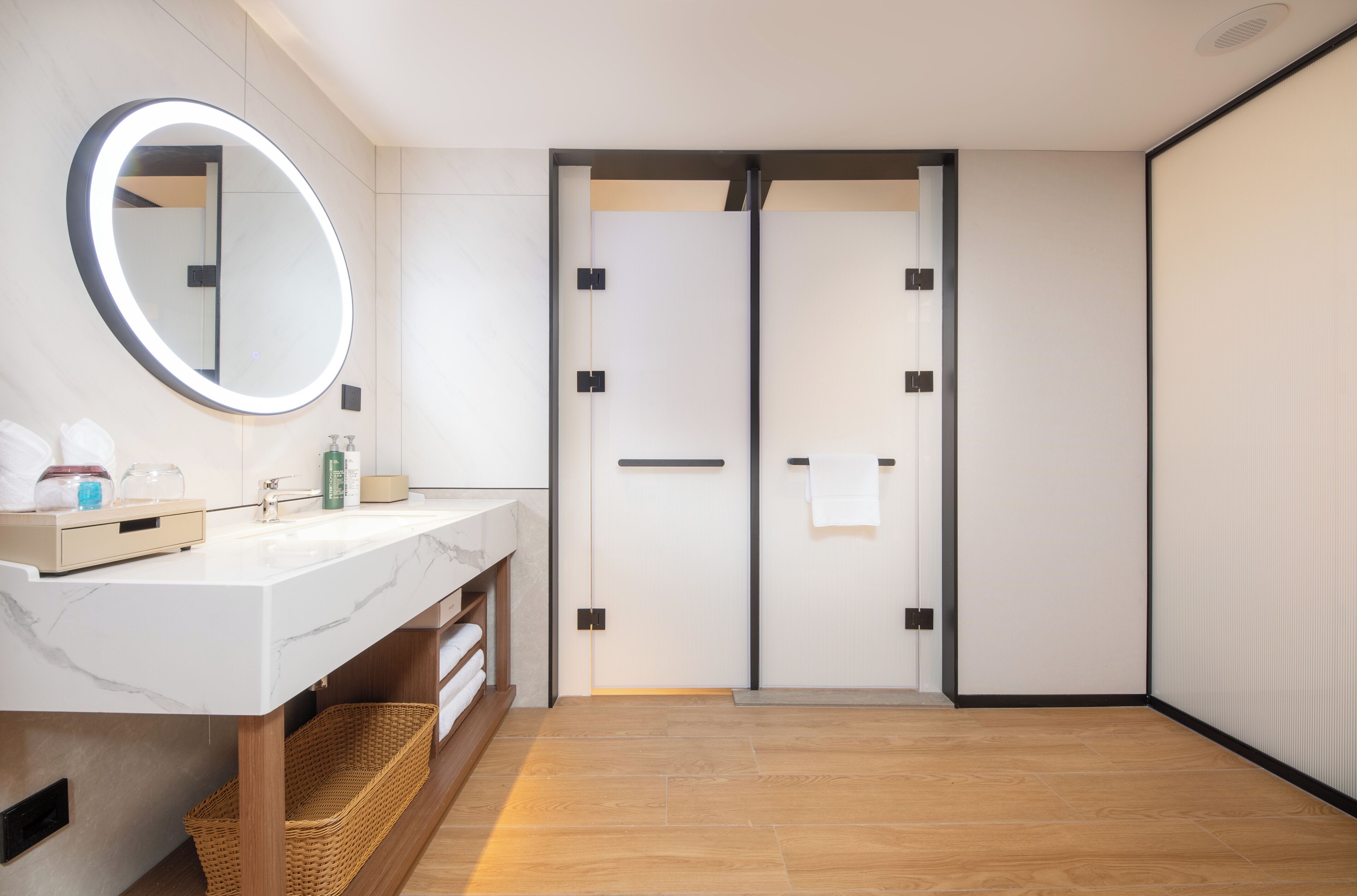 Un baño moderno con un lavamanos de mármol blanco, un espejo redondo con luz, suelo de madera y una puerta corredera.