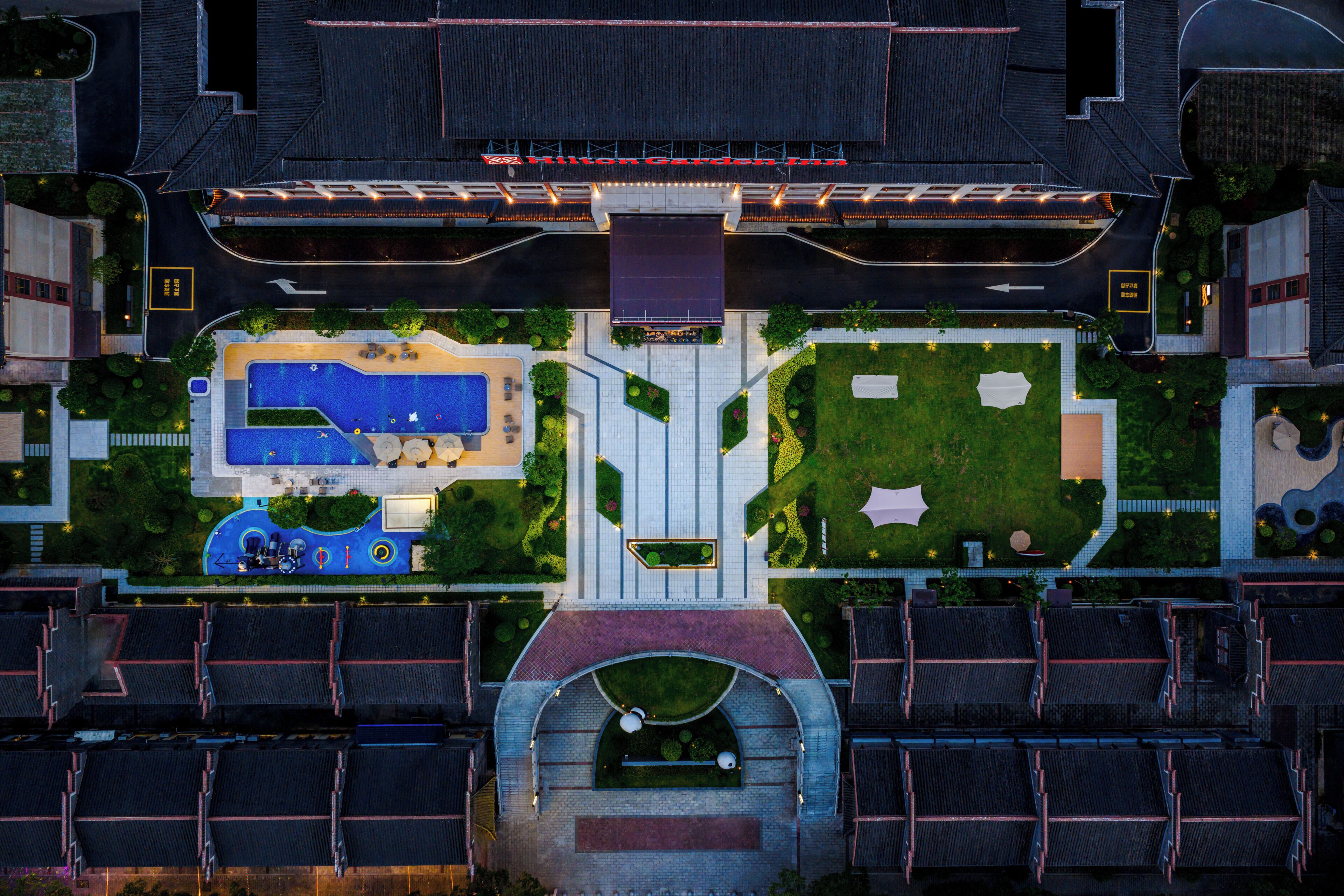 An aerial view of a hotel with a swimming pool, landscaped gardens, and a parking lot.