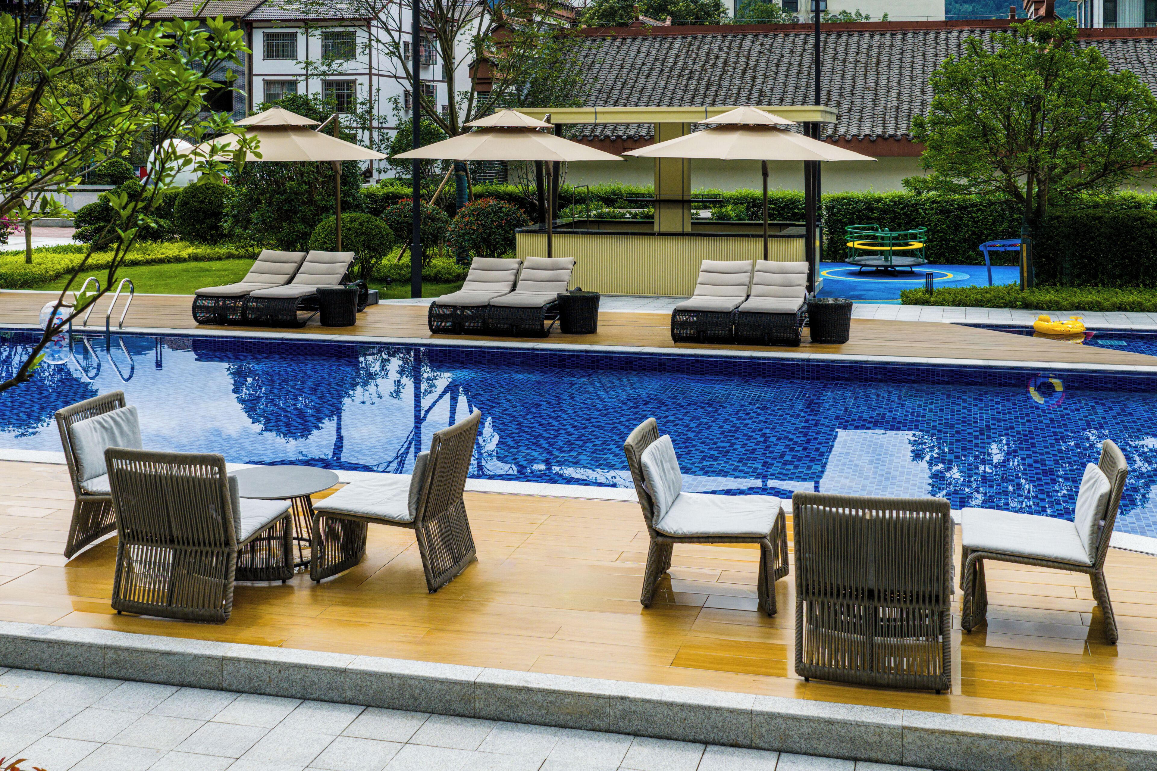 A pool area with lounge chairs and umbrellas, a wooden deck, and a view of buildings in the background.