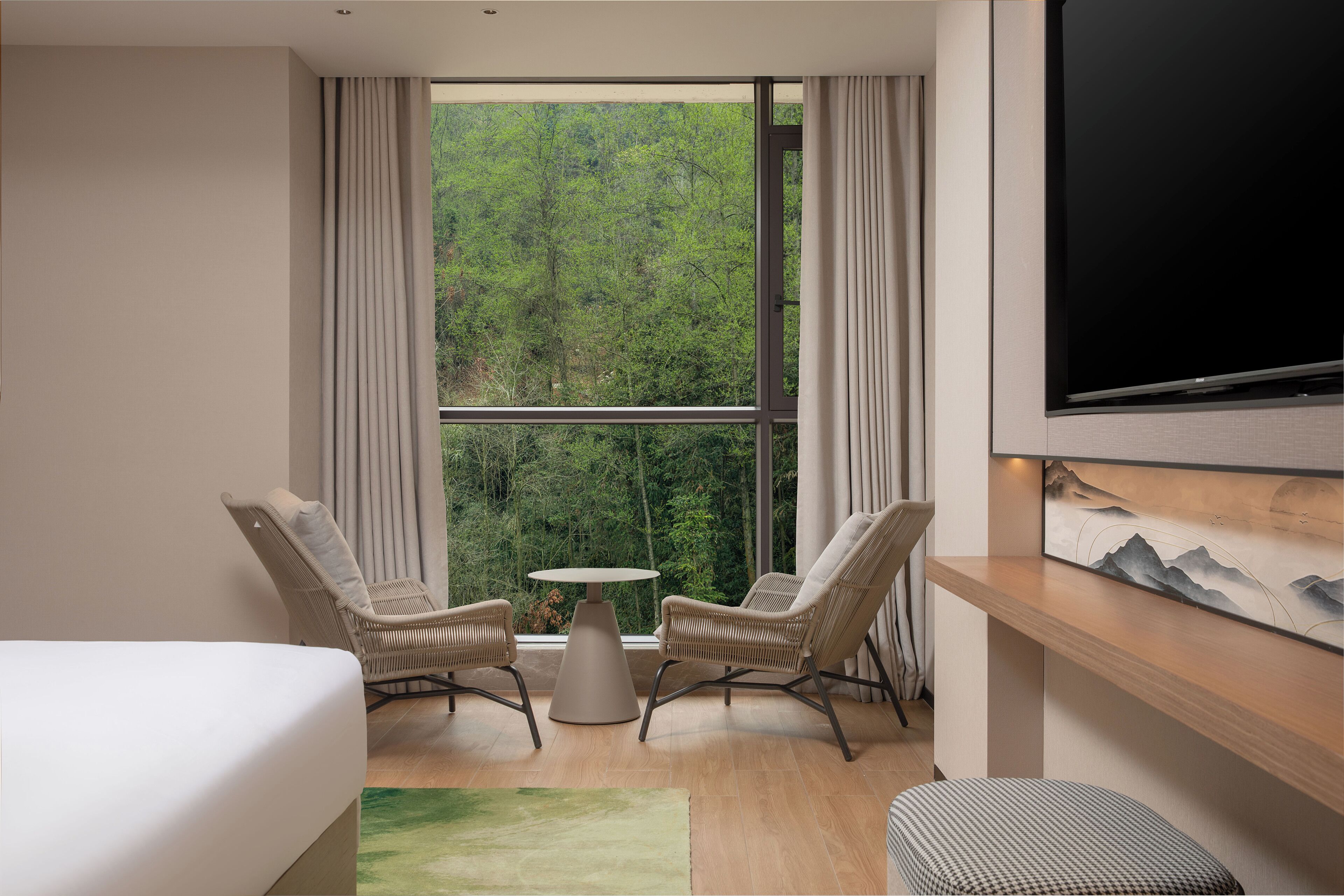 A room with a large window, two wicker chairs, a small round table, a bed, a TV, and a painting on the wall.
