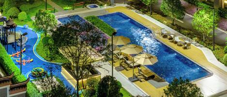 A large outdoor swimming pool with a children's play area, surrounded by well-maintained landscaping and modern buildings.