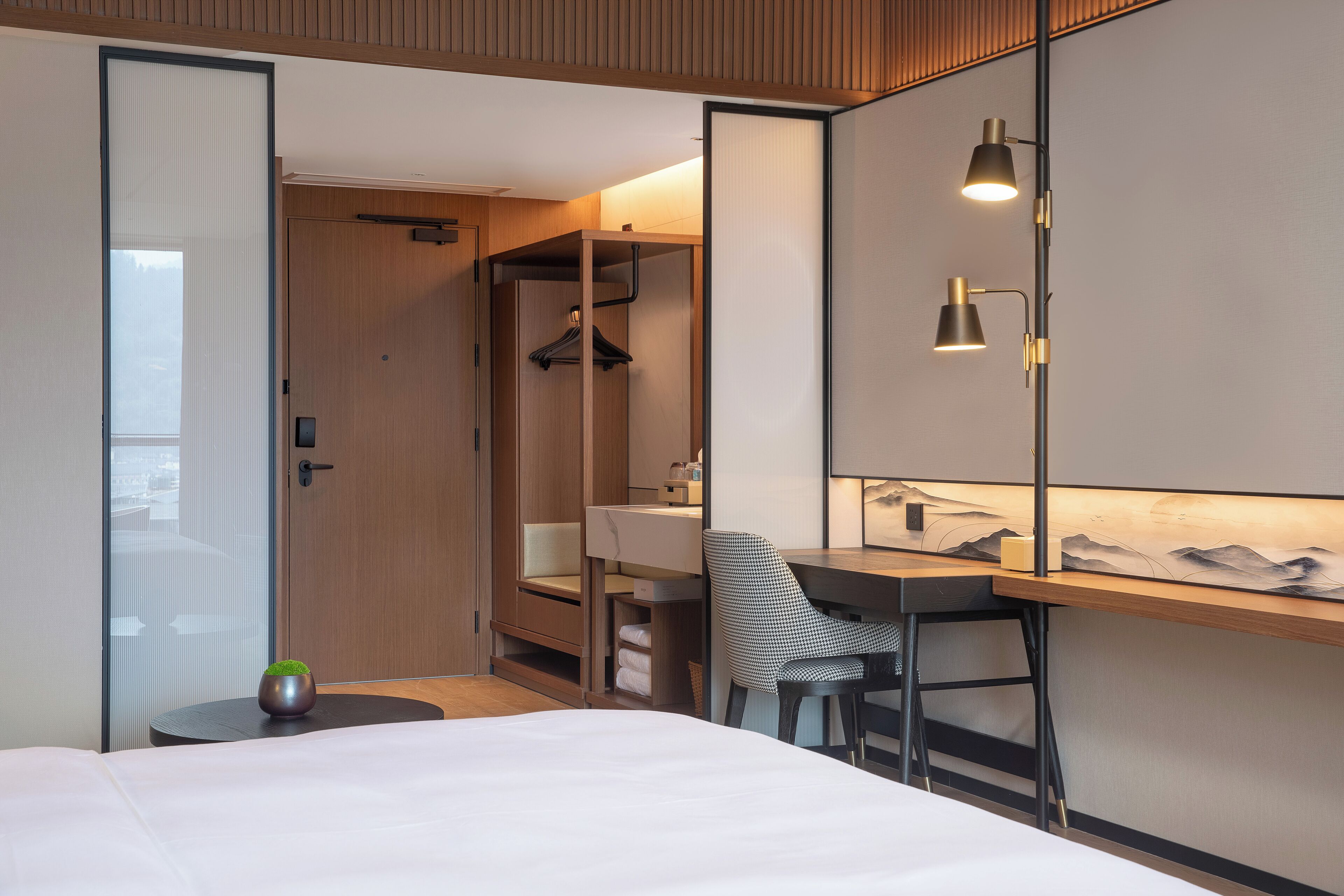 Una habitación de hotel moderna con una cama, un escritorio, una silla, una mesita con una planta y un armario con espejo.