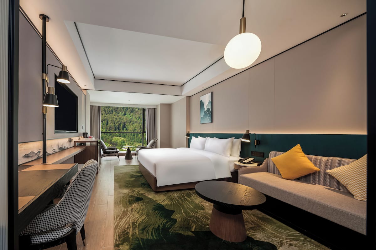 A modern hotel room with a large bed, a sofa, a dining table, and a view of a forest through a large window.