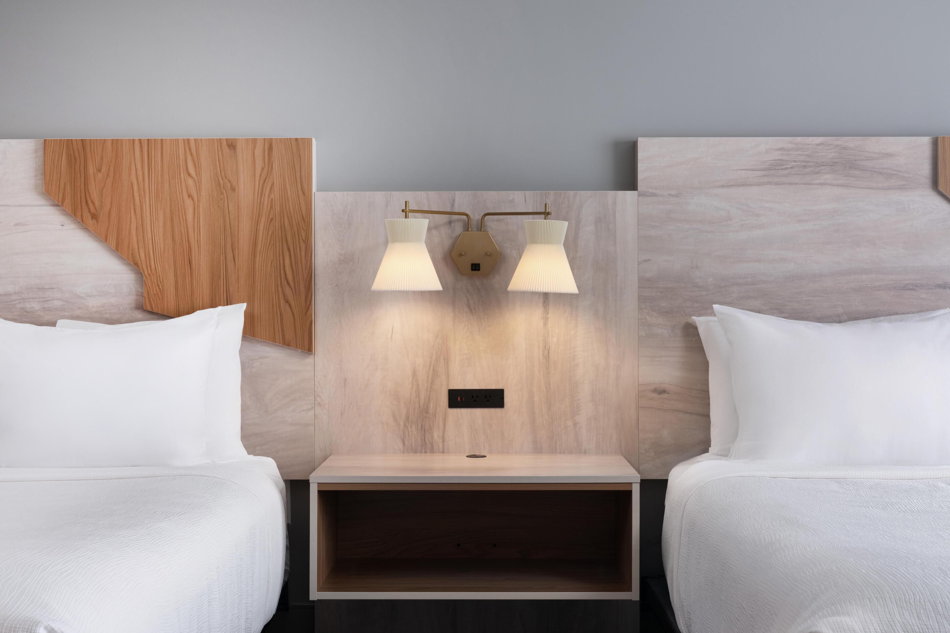 A hotel room with two beds, a wooden headboard, a nightstand, and a wall-mounted light fixture.