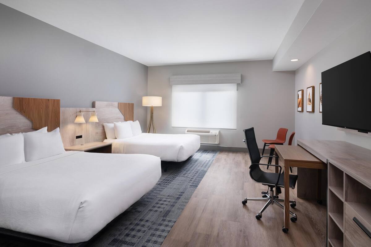 A hotel room with two beds, a desk, a chair, a TV, and a window with blinds.