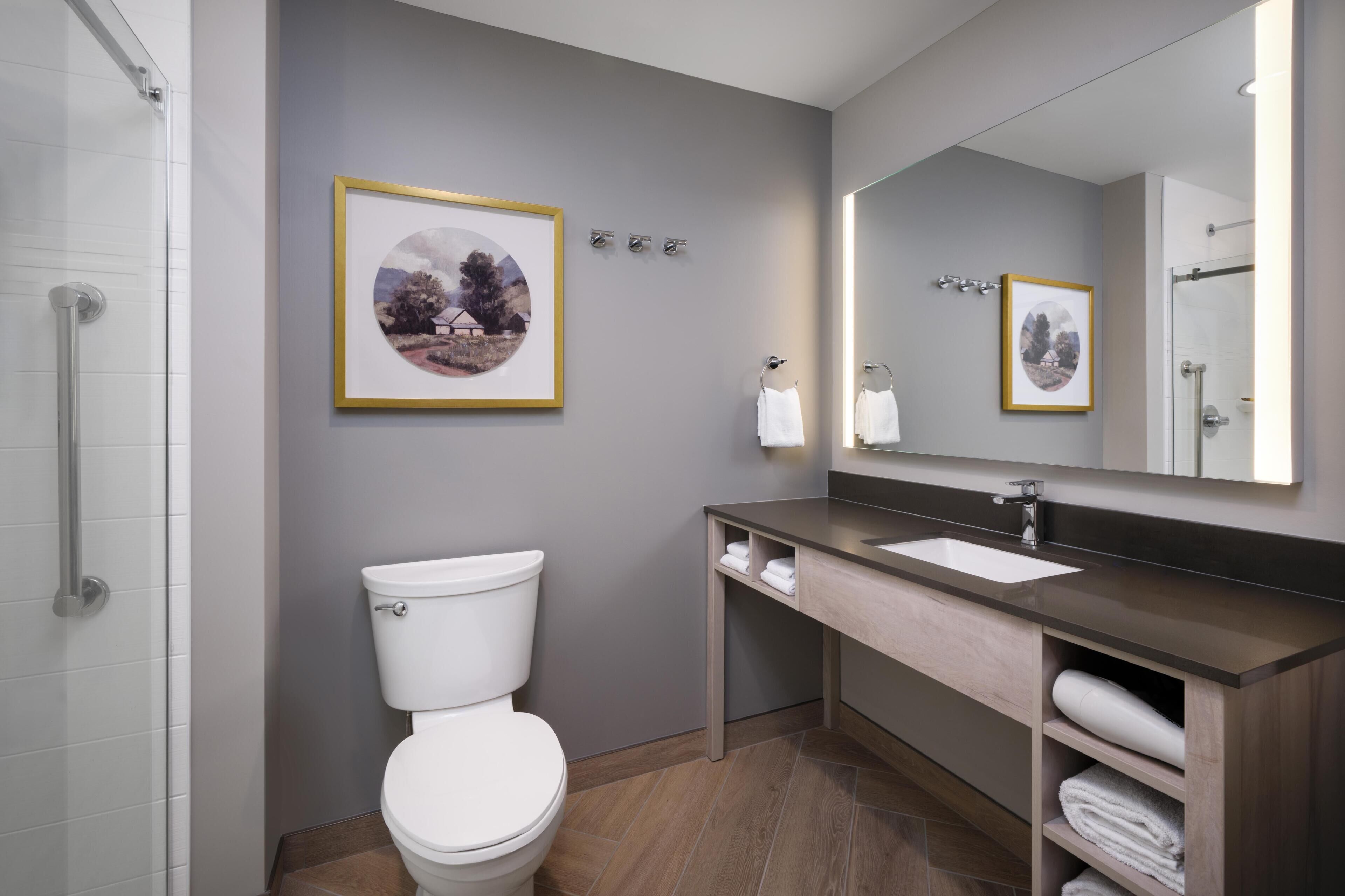 A modern bathroom with a toilet, sink, and mirror. There are framed pictures on the wall and a shower area.