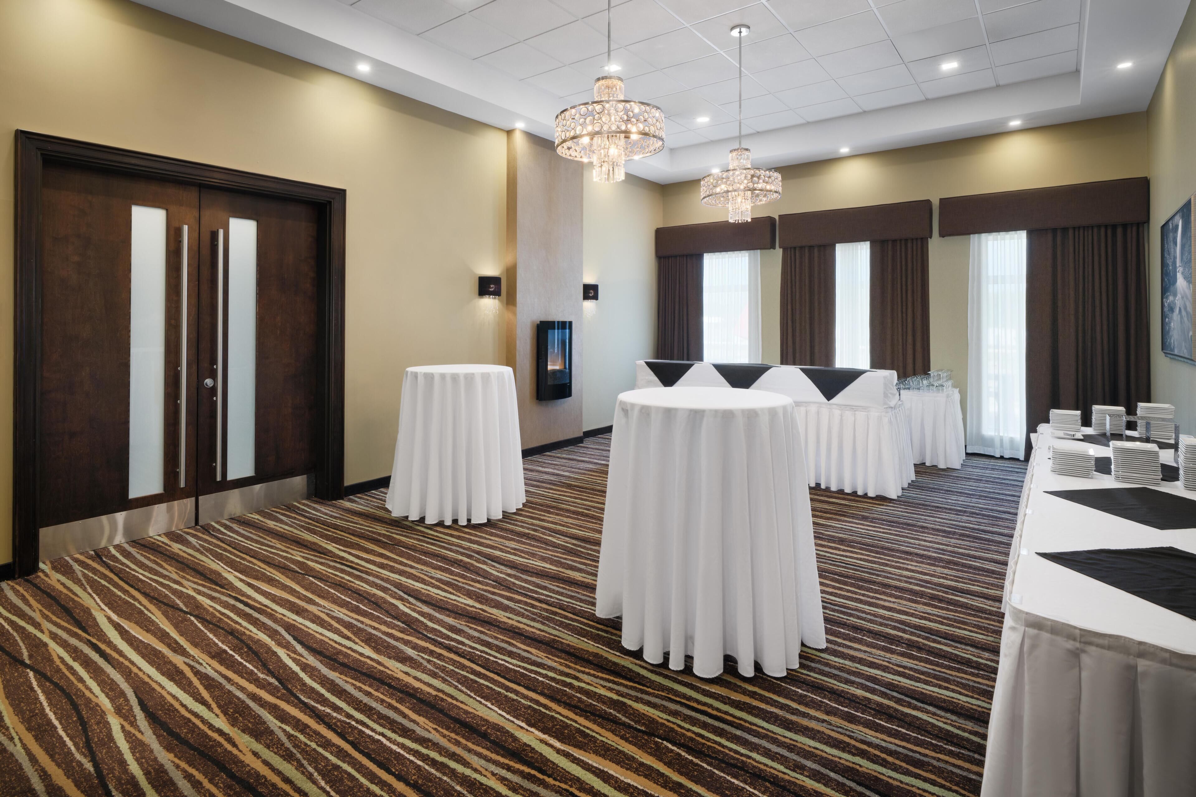 A hotel room with a large bed, a round table, a small round table, a minibar, and a large window with curtains.