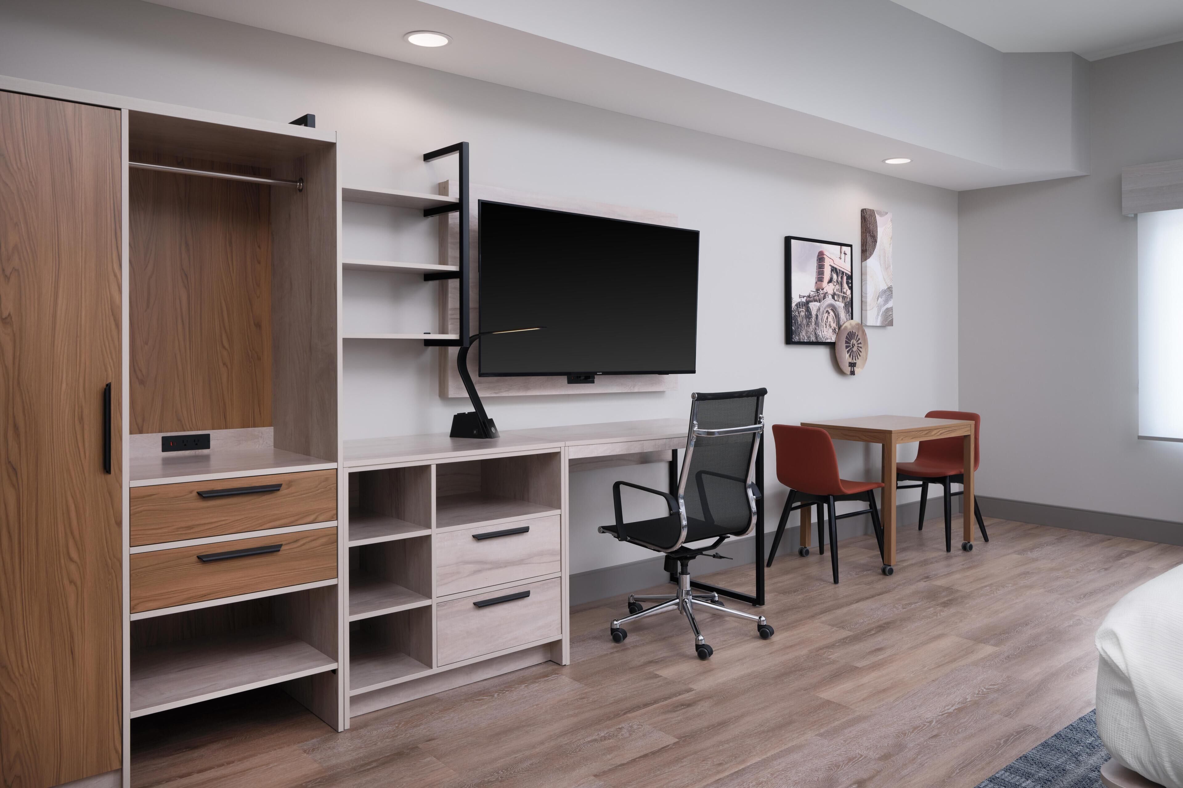 A modern room with a large flat-screen TV, a wooden wardrobe, a desk with a chair, and a dining area.