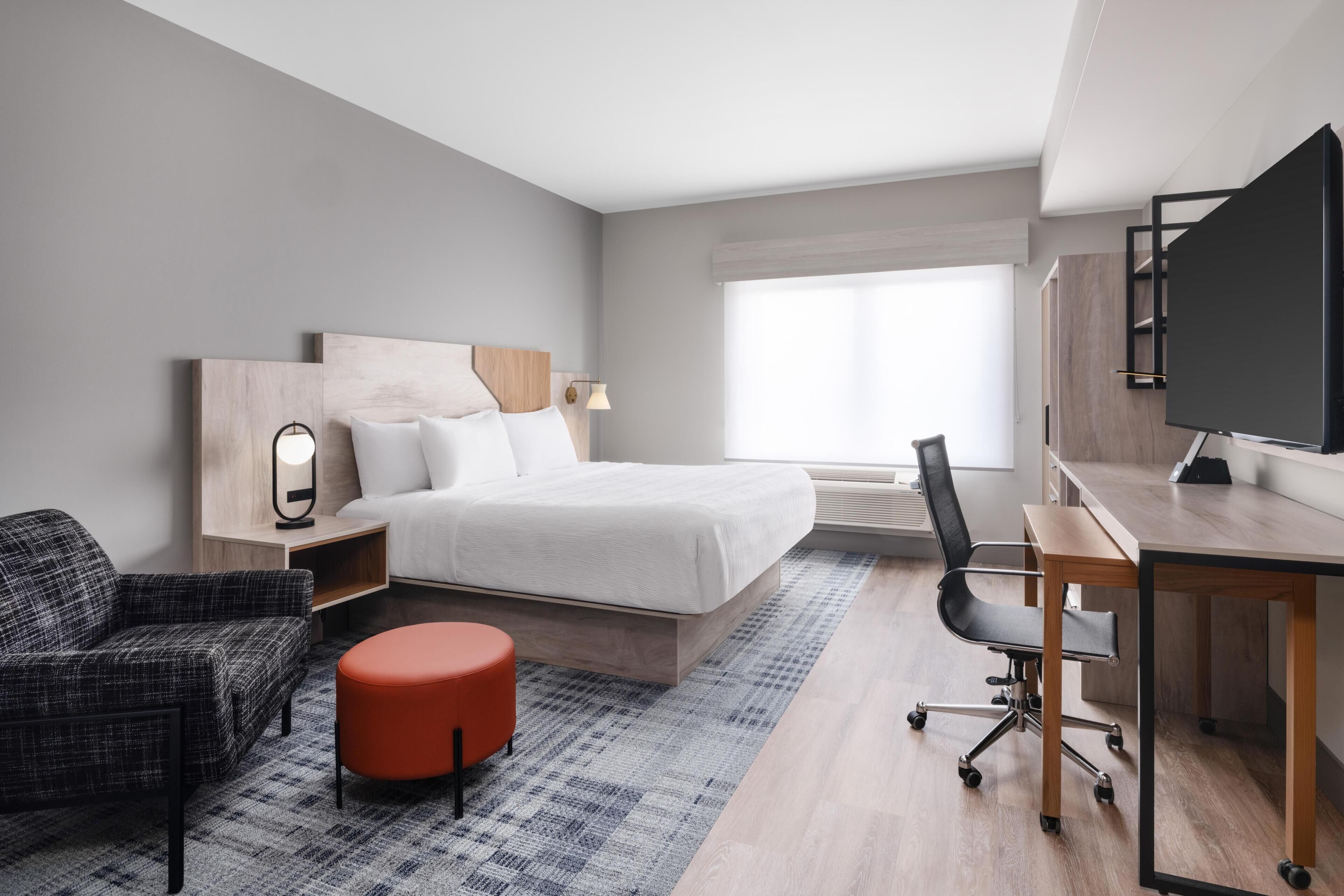 A modern hotel room with a bed, a desk, a chair, a red ottoman, and a television.