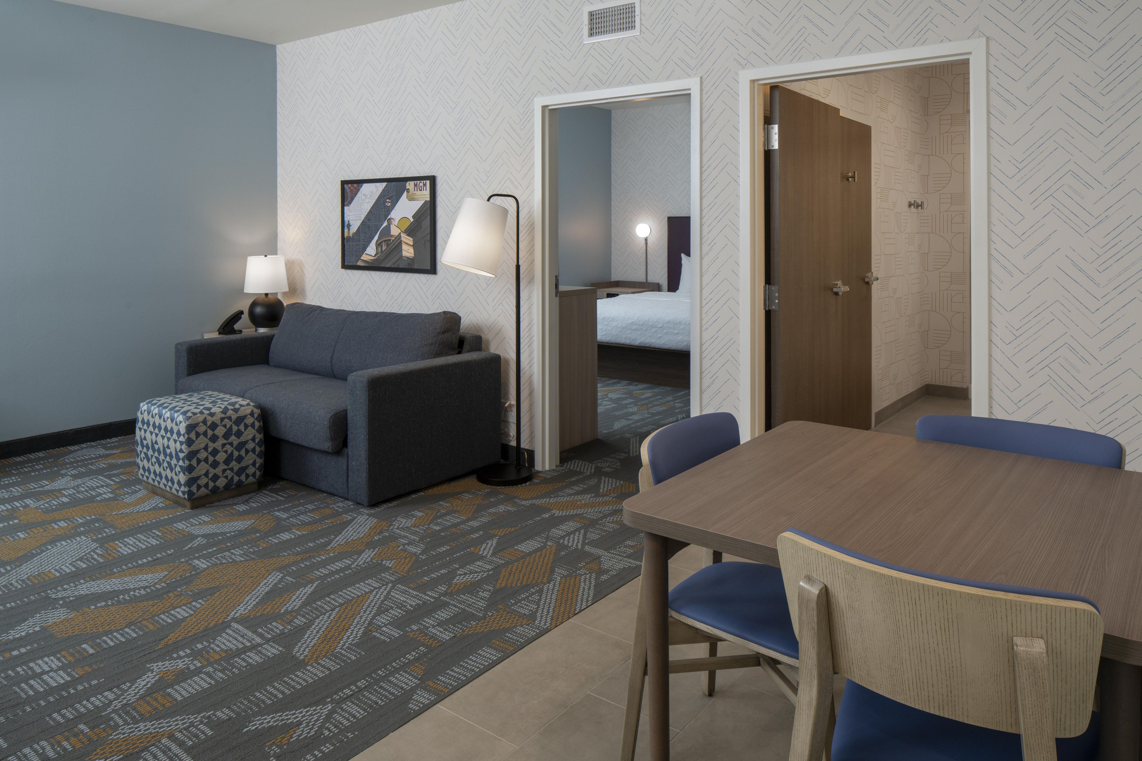A hotel room with a sofa, a small table, a lamp, a patterned rug, and a bed with a headboard.