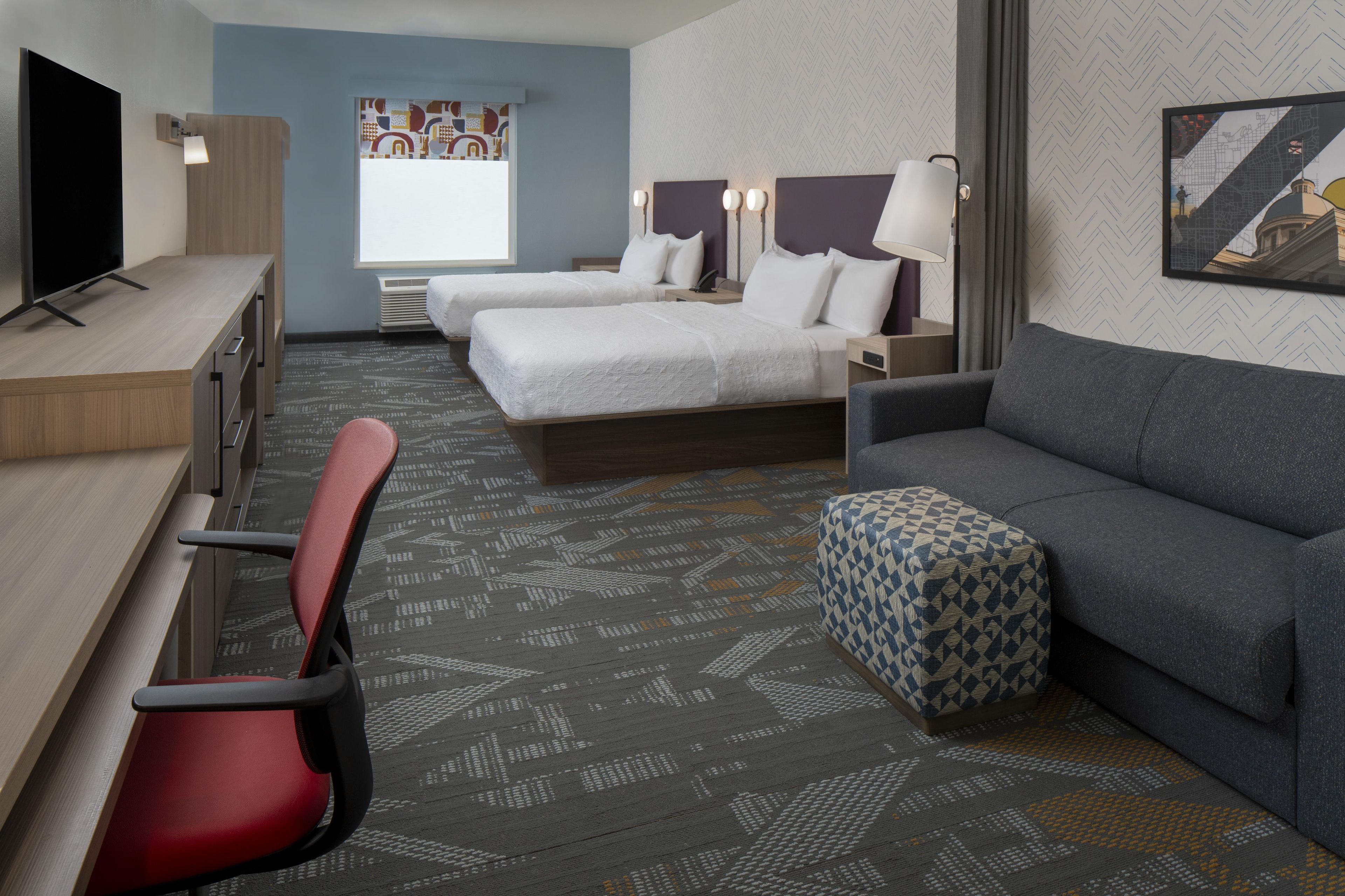 Una habitación de hotel con escritorio, dos camas, un sofá, una silla y alfombra estampada.