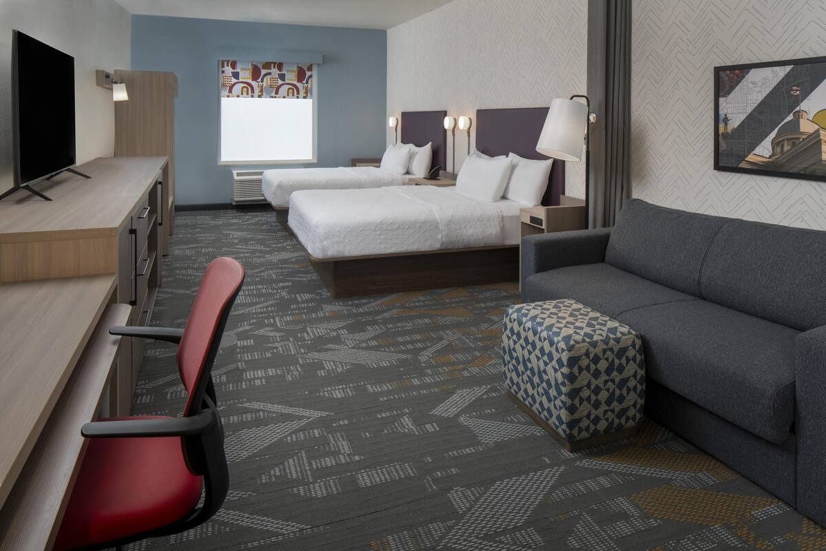Quarto de hotel com escrivaninha, duas camas, sofá, televisão e carpete estampado.