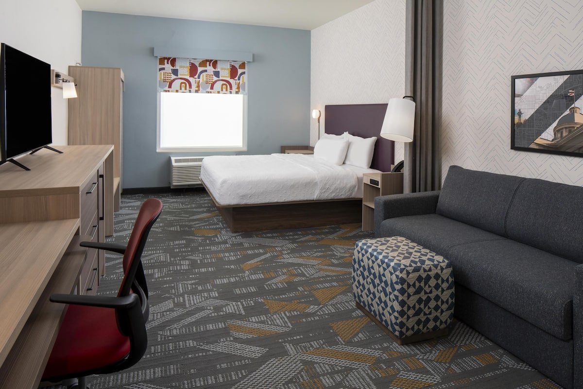 Quarto de hotel com cama, escrivaninha com televisão, sofá, carpete estampado e janela com vista.