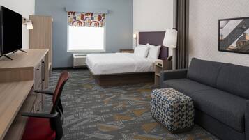 Habitación de hotel con cama, escritorio con televisor, sofá, alfombra estampada y ventana con vistas.