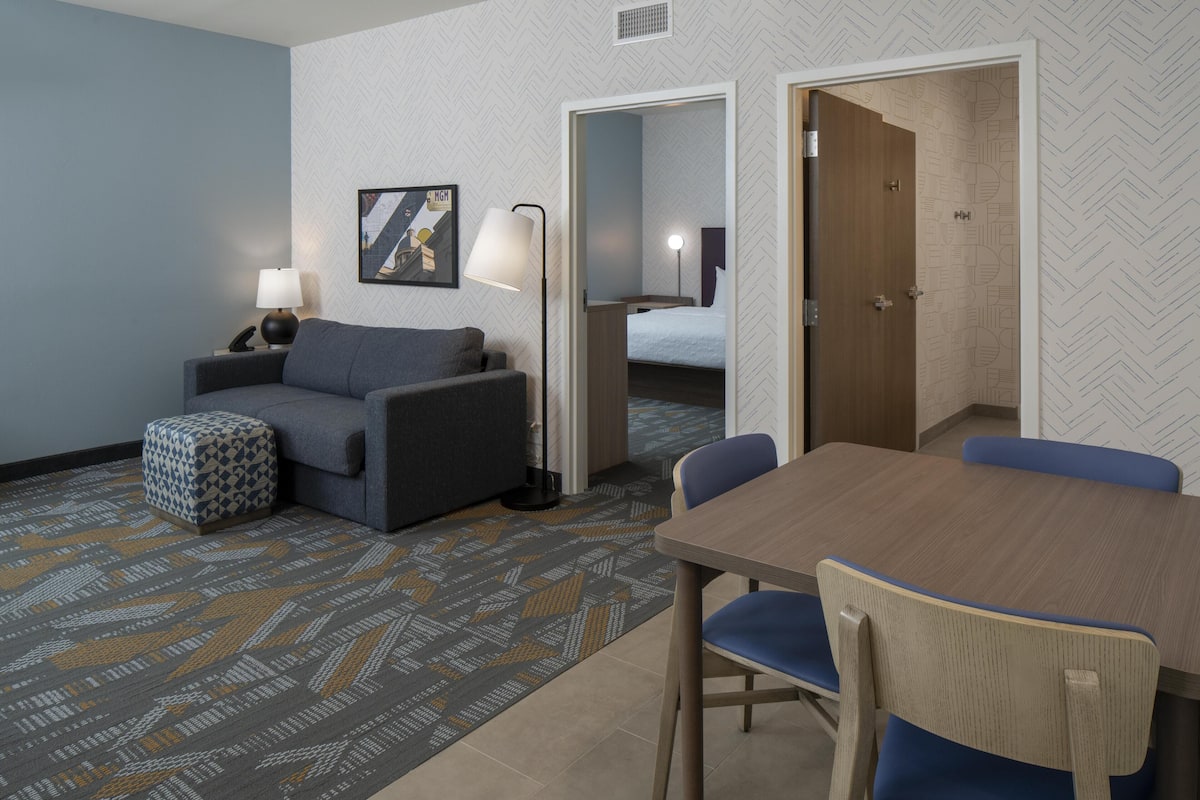 Quarto de hotel com sofá, um pequeno banquinho, mesa com cadeiras, luminária e cama ao fundo.