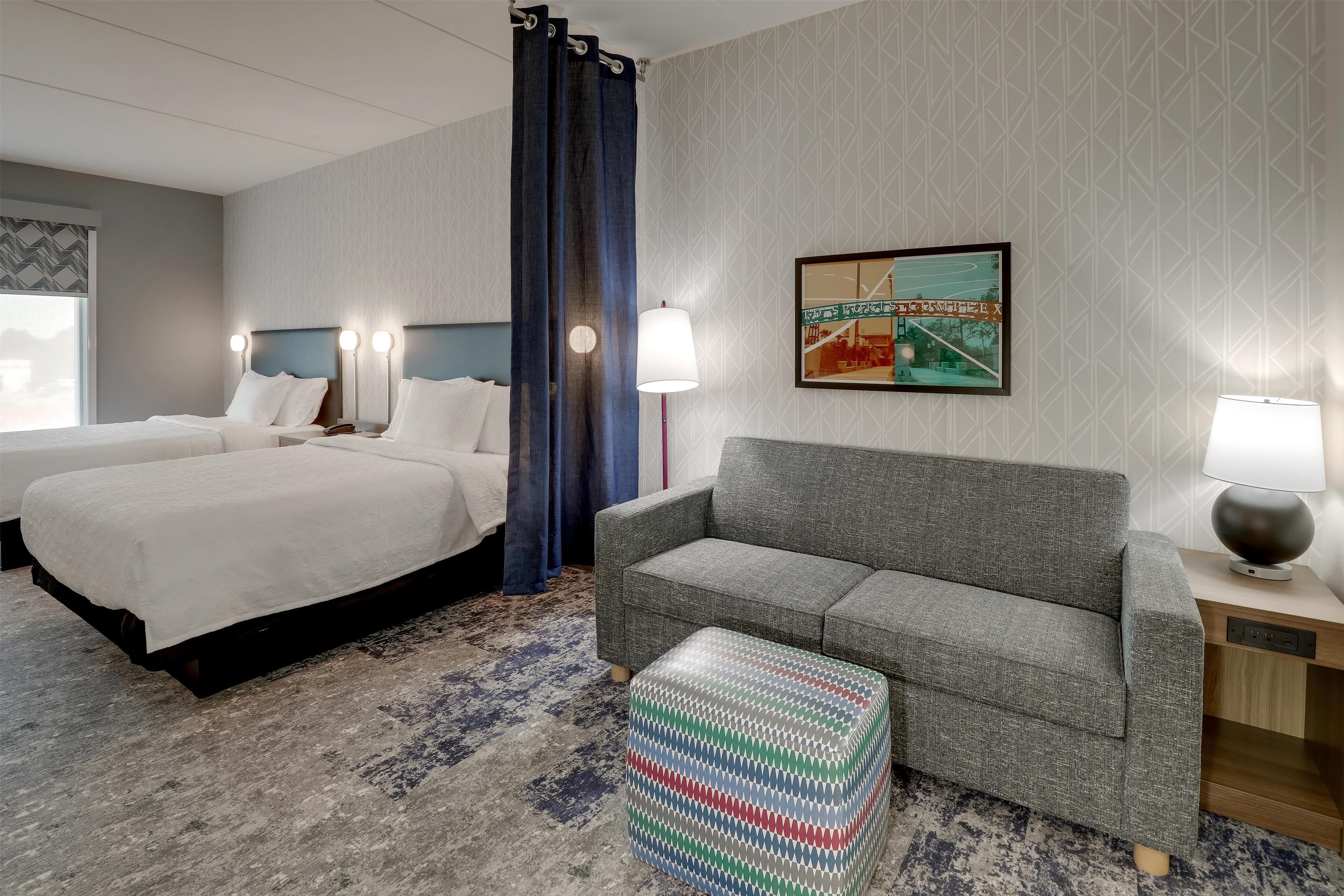 Habitación de hotel con dos camas, un sofá gris, un otomano estampado y un cuadro en la pared.