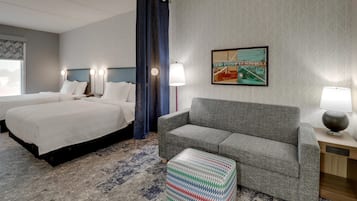 Habitación de hotel con dos camas, un sofá gris, un otomano estampado y un cuadro en la pared.