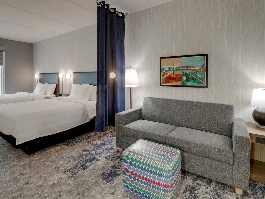 Habitación de hotel con dos camas, un sofá gris, un otomano estampado y un cuadro en la pared.
