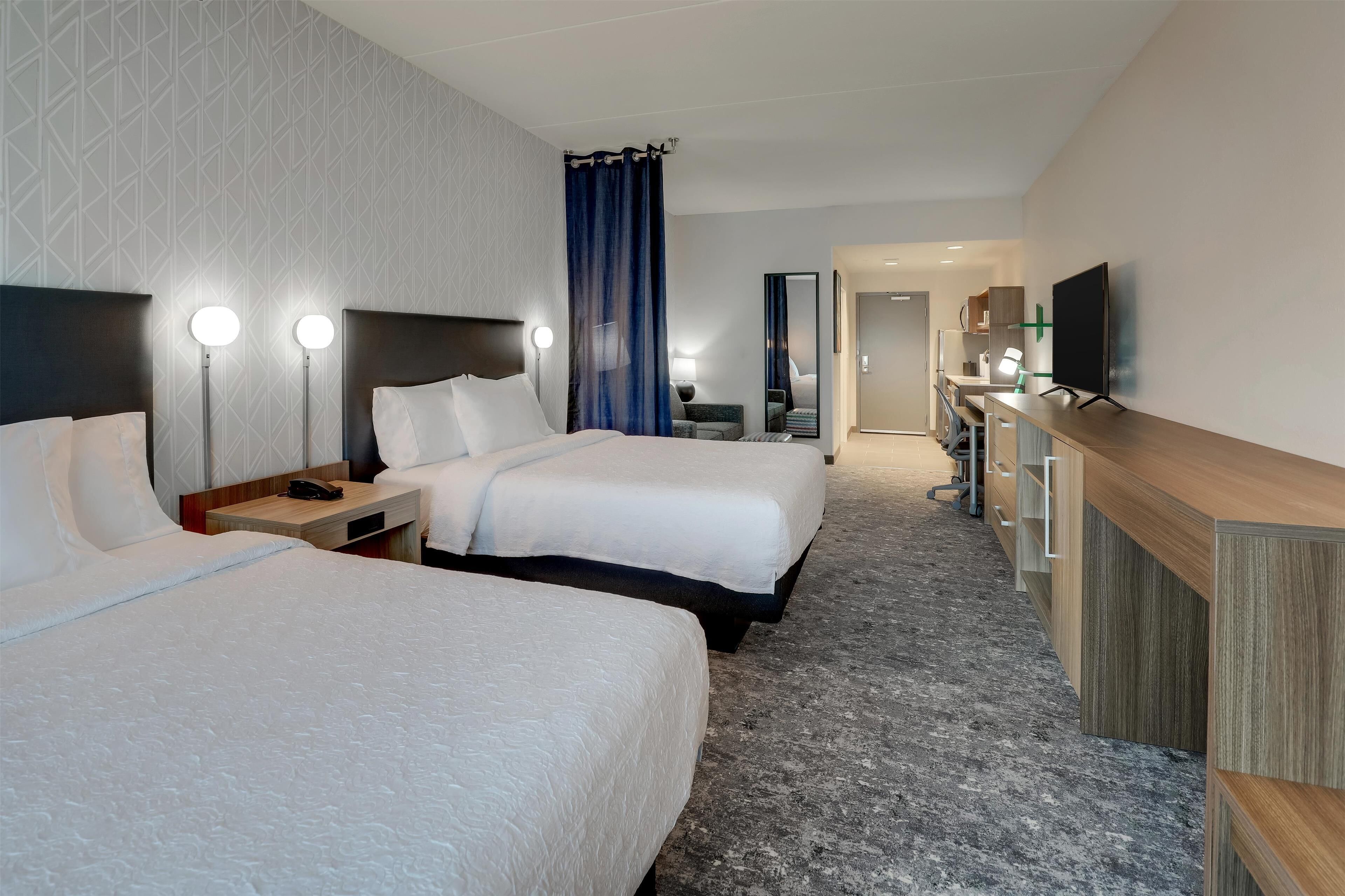 A hotel room with two beds, a desk, a TV, and a bathroom visible in the background.