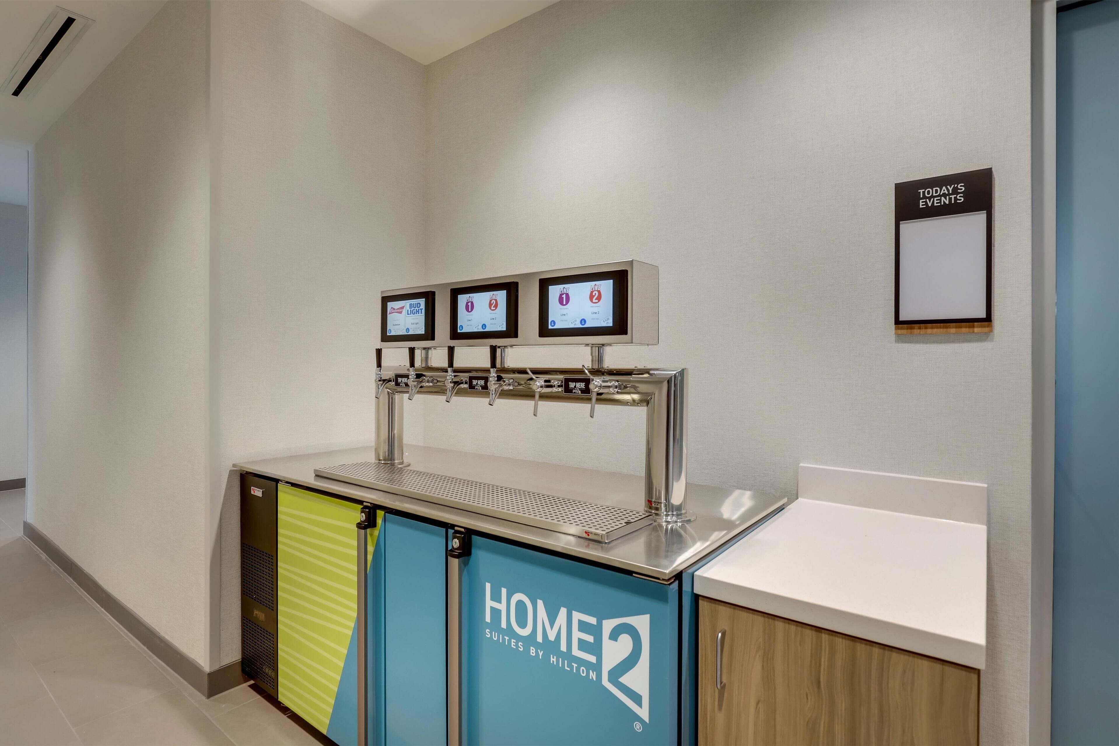Zona de recepció d'hotel amb una estació de begudes autoserveïda, un cartell amb els actes del dia i un taulell amb un cartell de 'HOME 2 Suites by Hilton'.