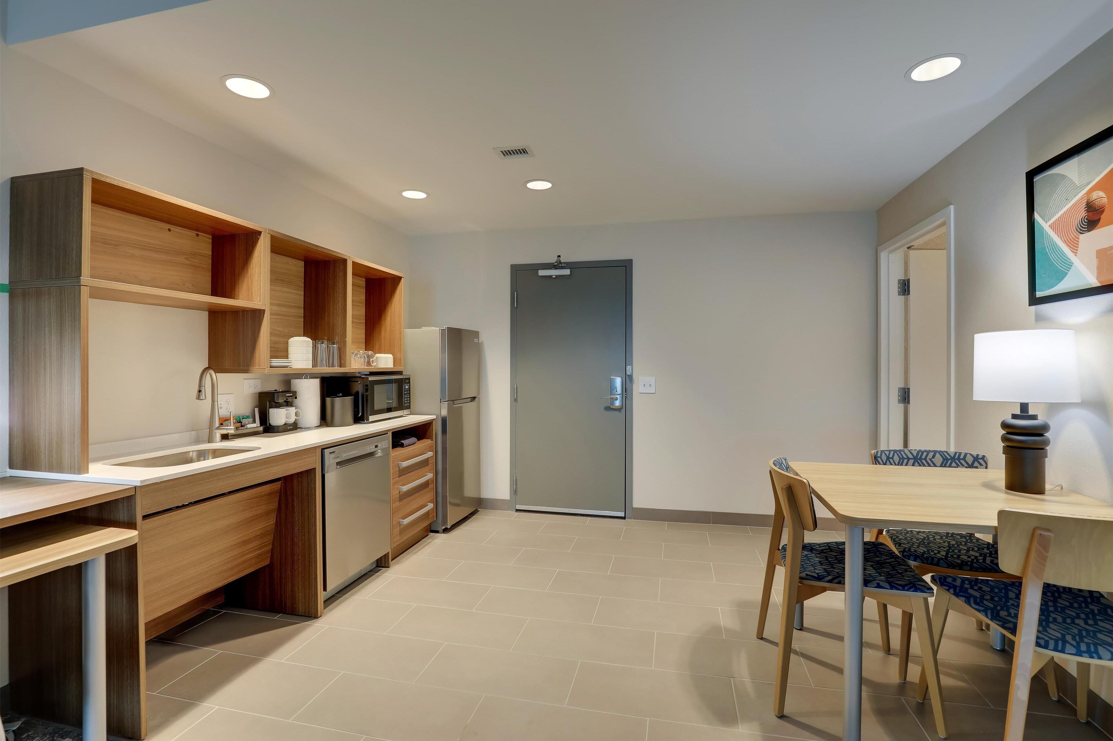 A modern kitchen with wooden cabinetry, a built-in microwave, and a coffee machine. There is a dining area with a table and chairs, and a door leading to another room.