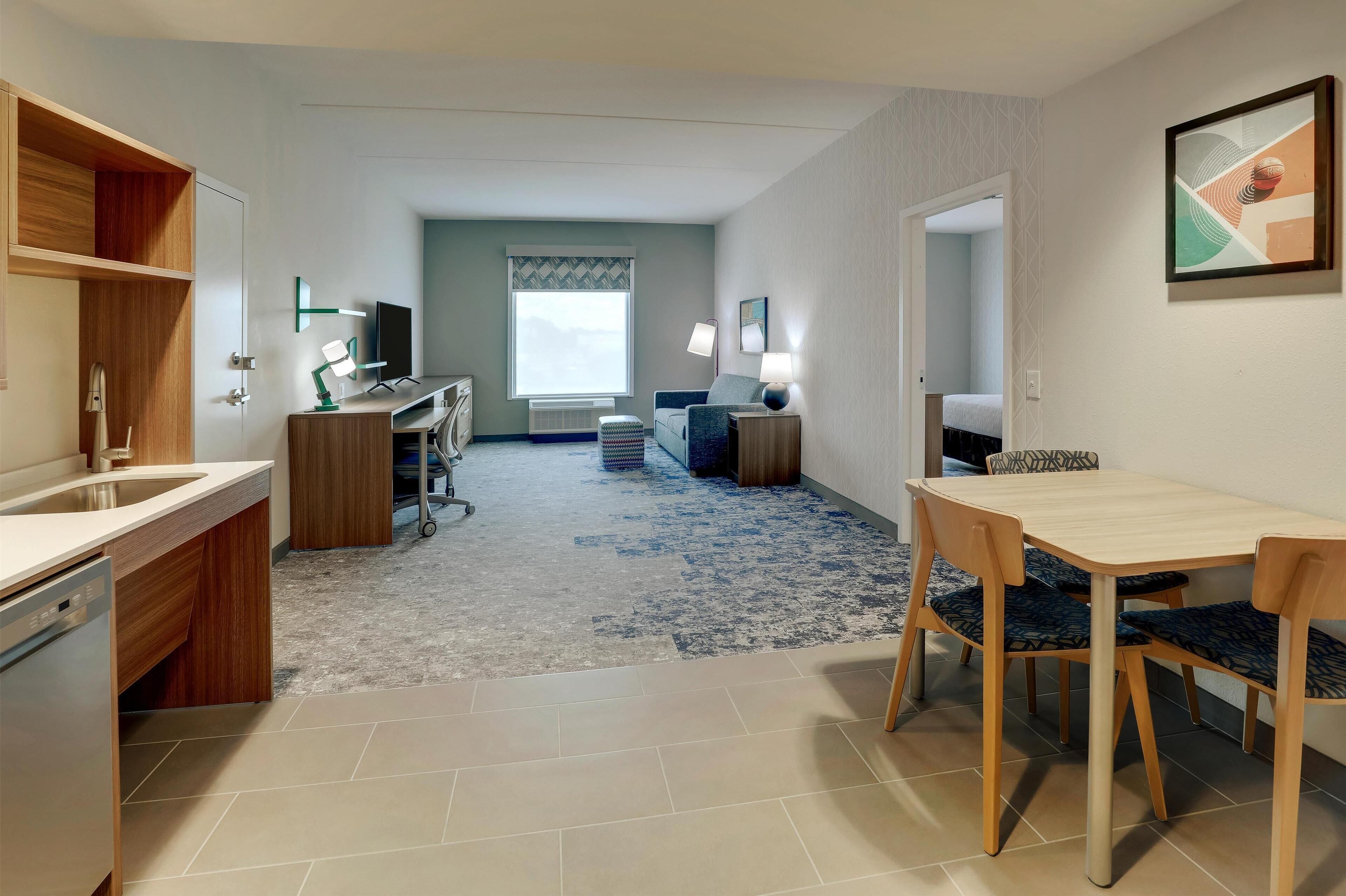 A modern hotel room with a kitchen area, a dining table with chairs, a sofa, a TV, and a large window with a view.