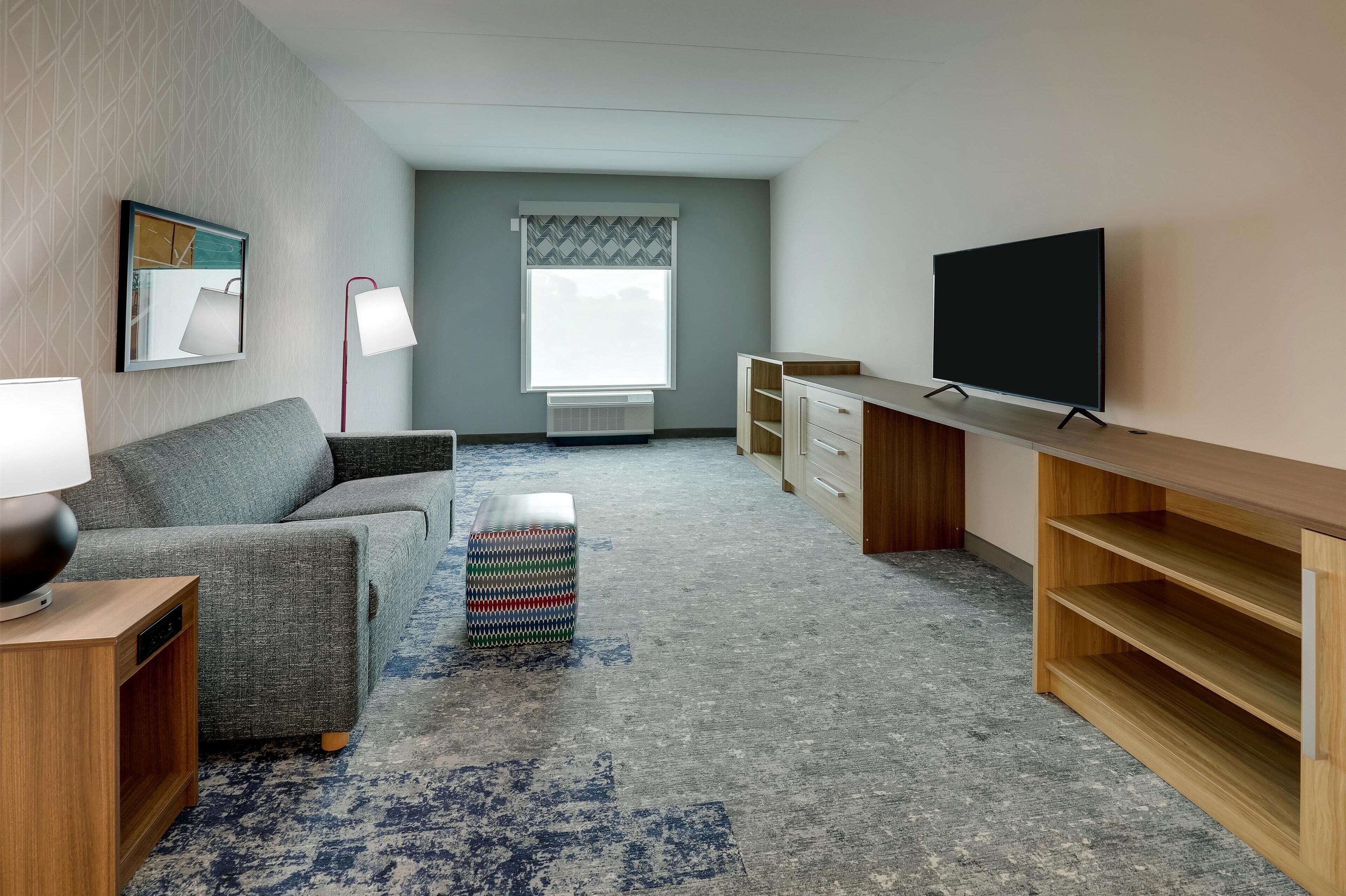 A modern living room with a grey sofa, a small ottoman, a TV on a wooden stand, and a window with a patterned curtain.