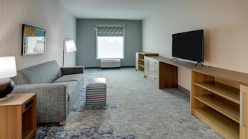 A modern living room with a grey sofa, a small ottoman, a TV on a wooden stand, and a window with a patterned curtain.