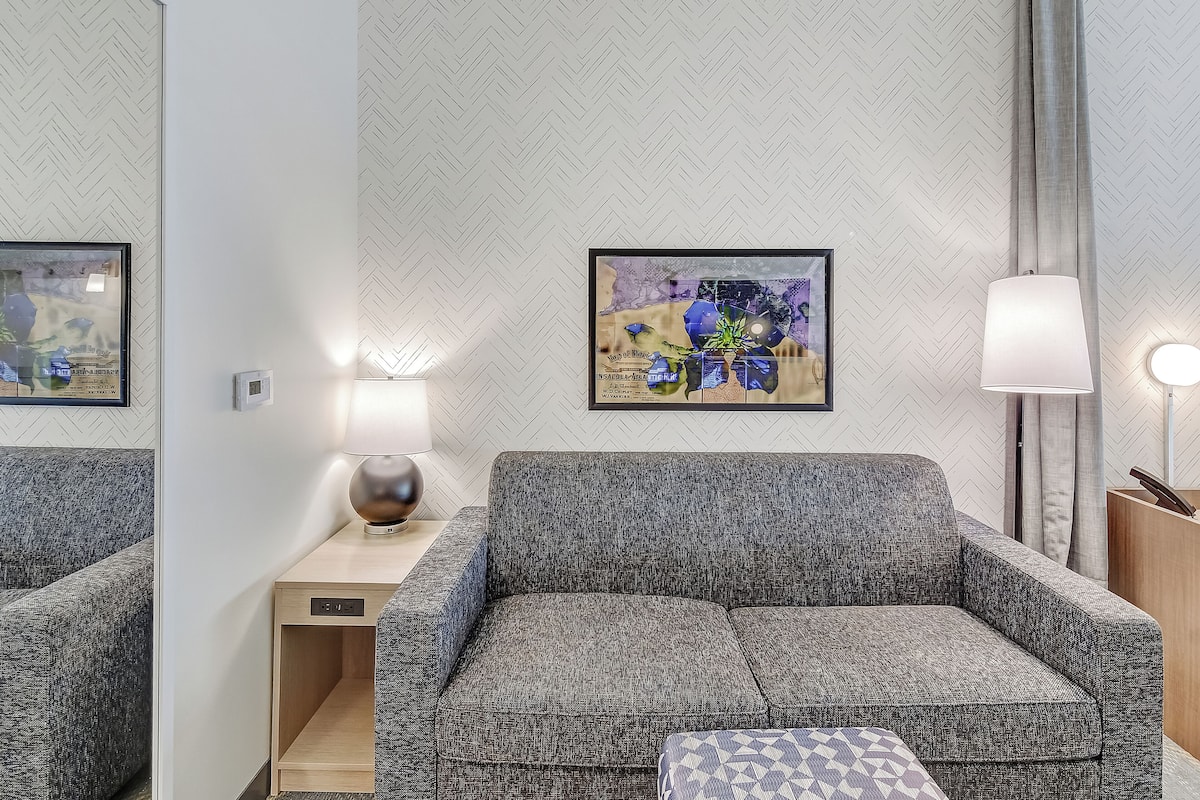 Une chambre d’hôtel moderne avec un canapé gris, une petite table d’appoint en bois, une lampe et une œuvre d’art encadrée au mur.