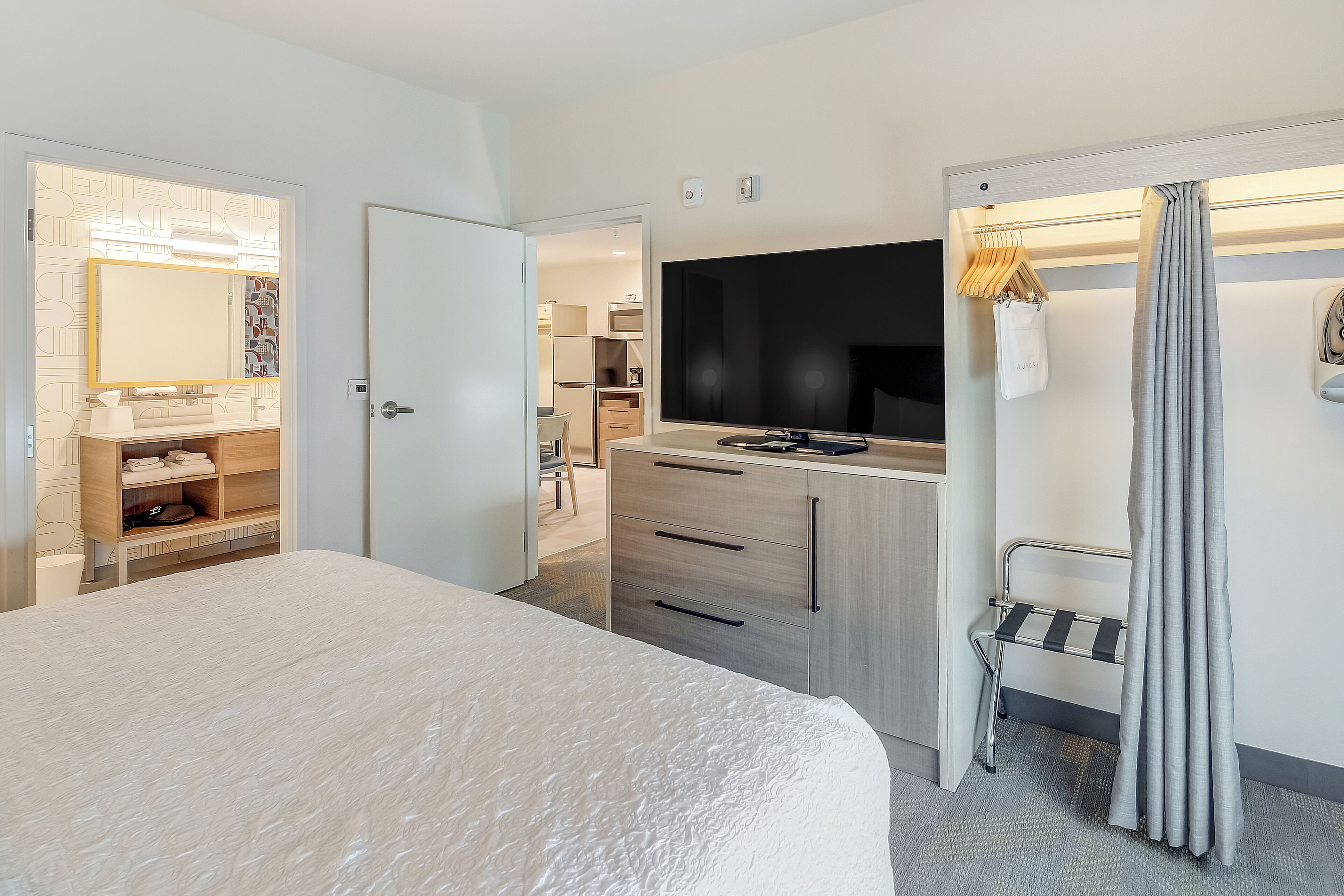 A modern hotel room with a large bed, a TV, a wardrobe, and a bathroom visible through an open door.