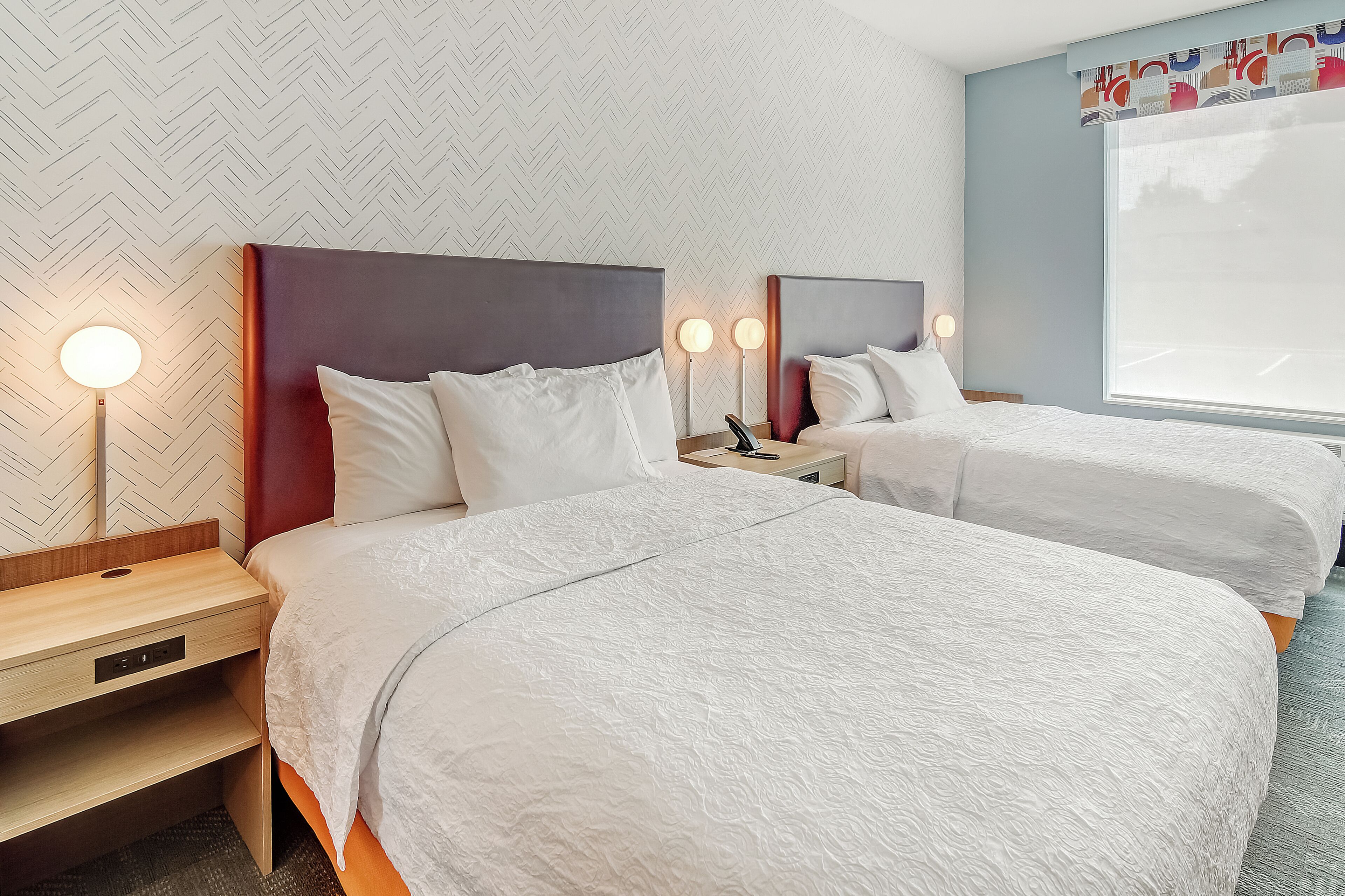 A hotel room with two beds, a wooden nightstand, a desk, and a large window with a view.