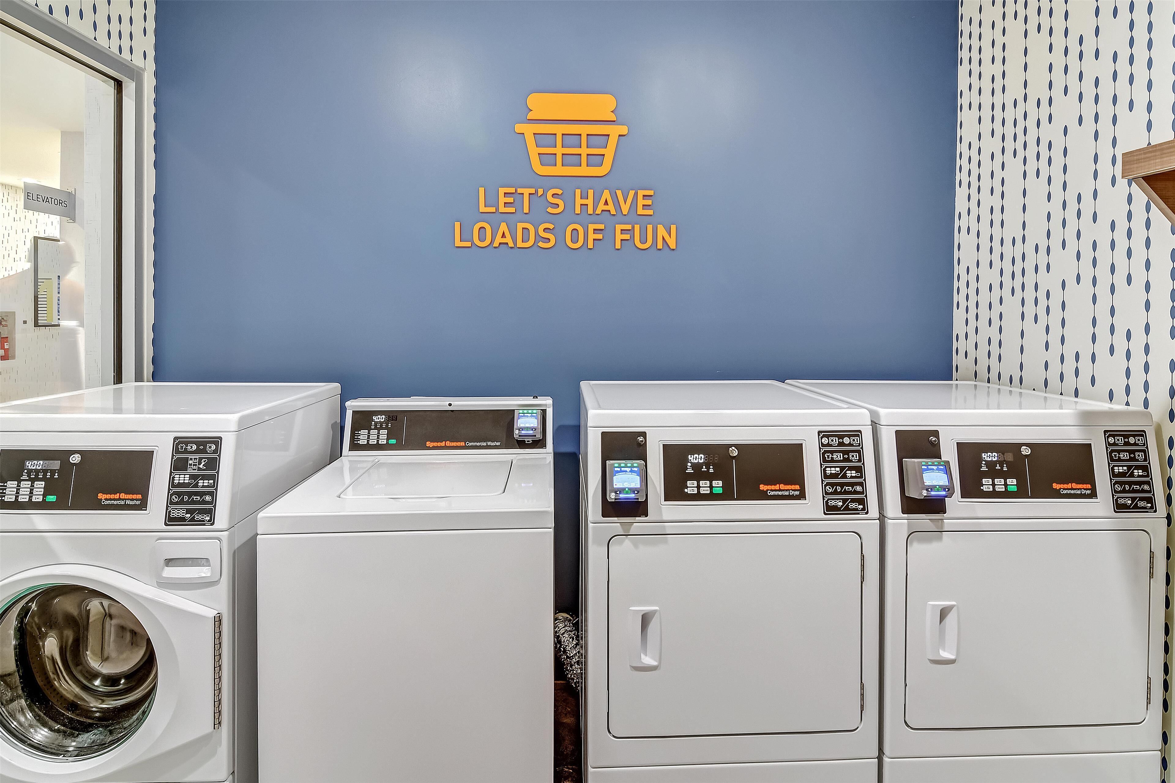 A laundromat with a blue wall and a sign that reads 'LET'S HAVE LOADS OF FUN'. There are three washing machines and two dryers.