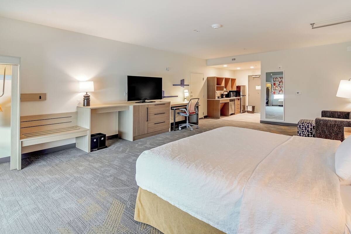 Une chambre d’hôtel avec un grand lit, un bureau avec une télévision, une chaise, un banc et une armoire.