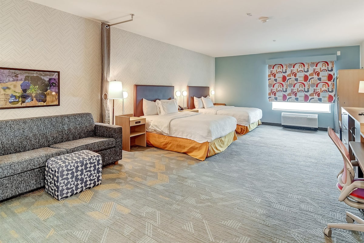 Une chambre d’hôtel avec un canapé, deux lits, un pouf à motifs, un bureau et une fenêtre avec des rideaux.