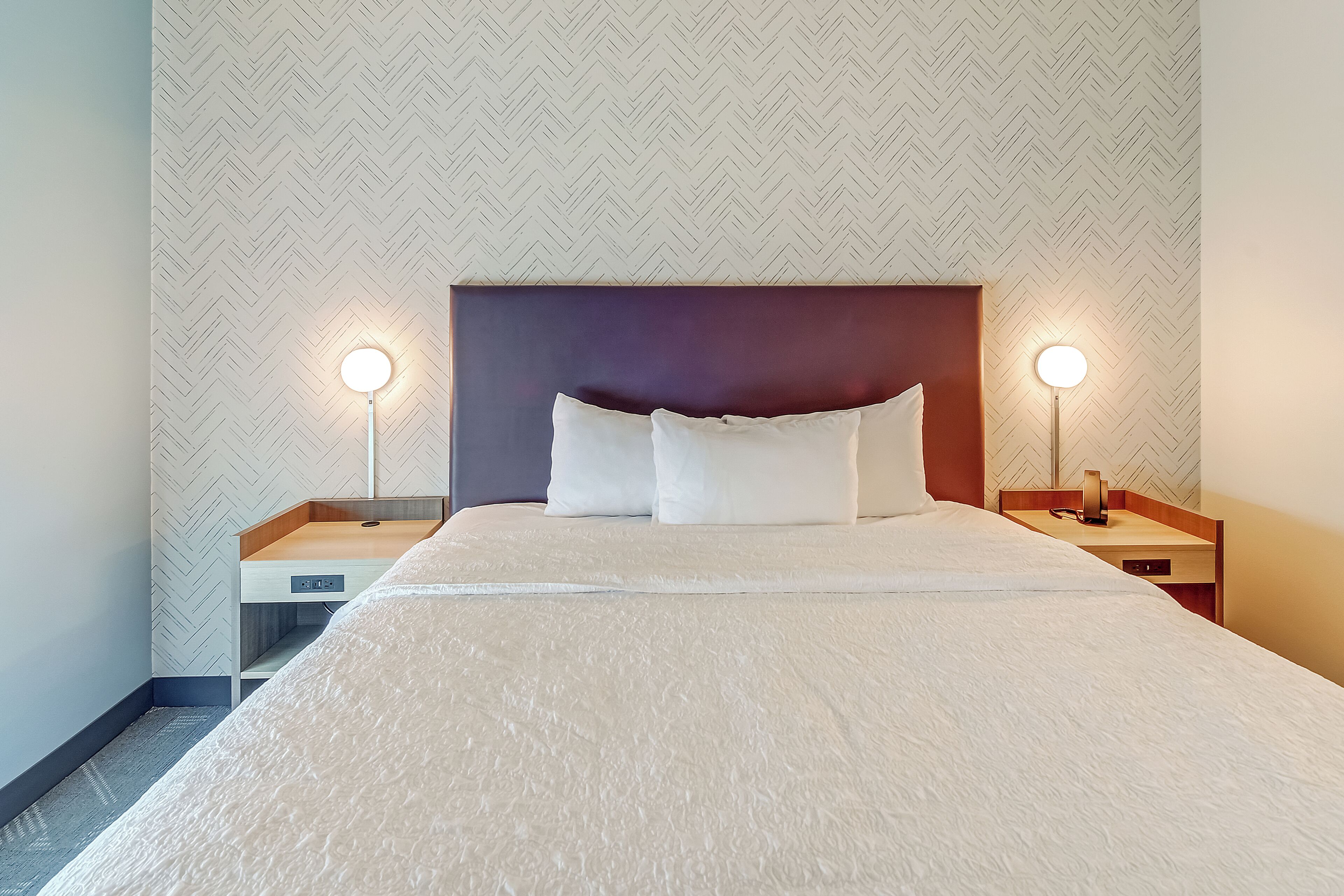 A neatly made bed with a white duvet, two bedside lamps, and a herringbone-patterned wall.