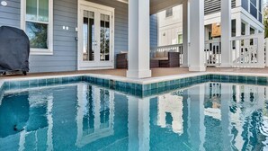 En swimmingpool med klart blåt vand, omgivet af en overdækket terrasse med hvide søjler og udsigt til et hus i baggrunden.
