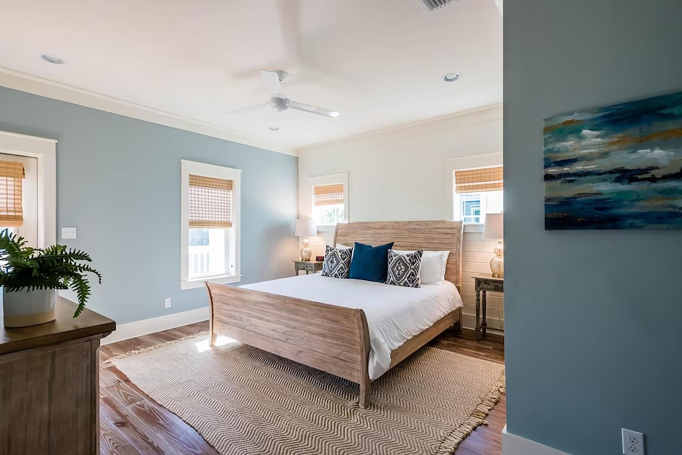A bedroom with a wooden bed, a large window with blinds, a ceiling fan, and a painting on the wall.