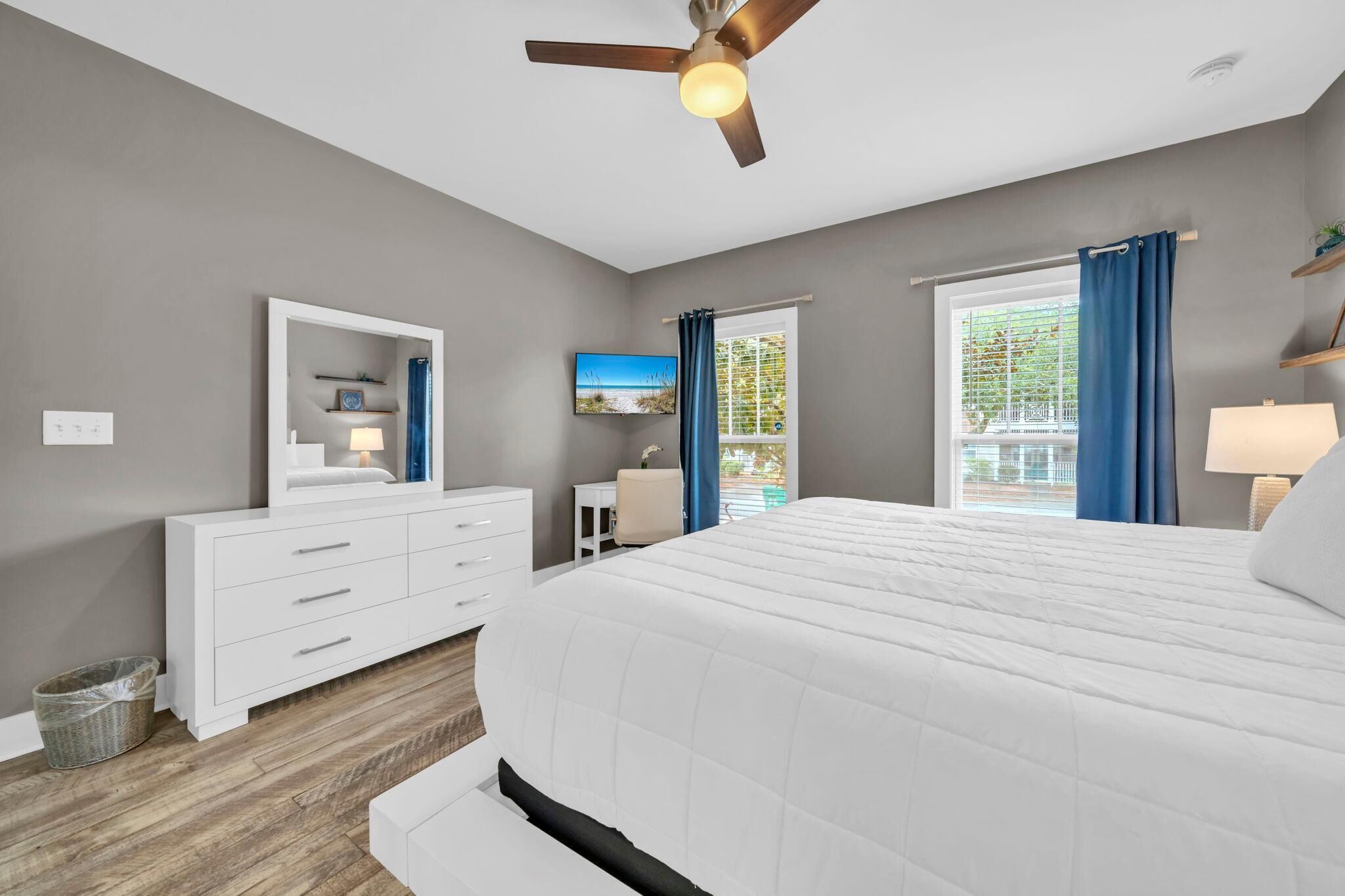 Quarto com cama grande, ventoinha no teto, escrivaninha e janela com cortinas.