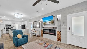 A modern living room with a flat-screen TV, a fireplace, and a dining area with a blue armchair and a wooden coffee table. - Kitties Koop (Santa Rosa Beach)